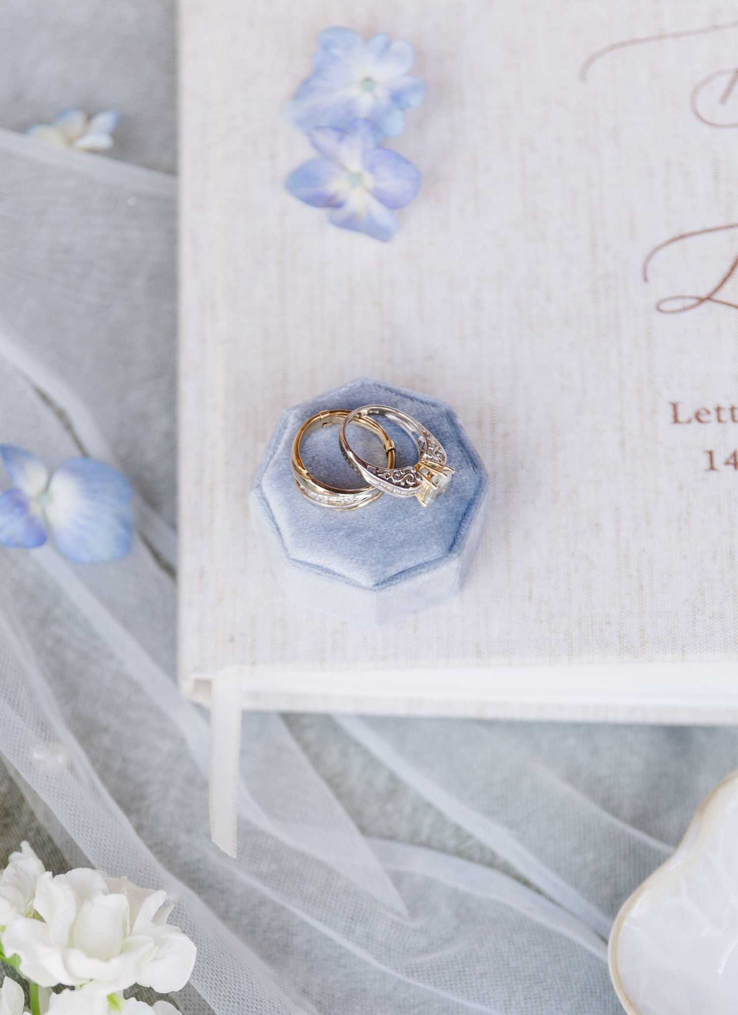 Two gold wedding bands in a blue velvet ring box atop a wedding guest book with hydrangea blooms.