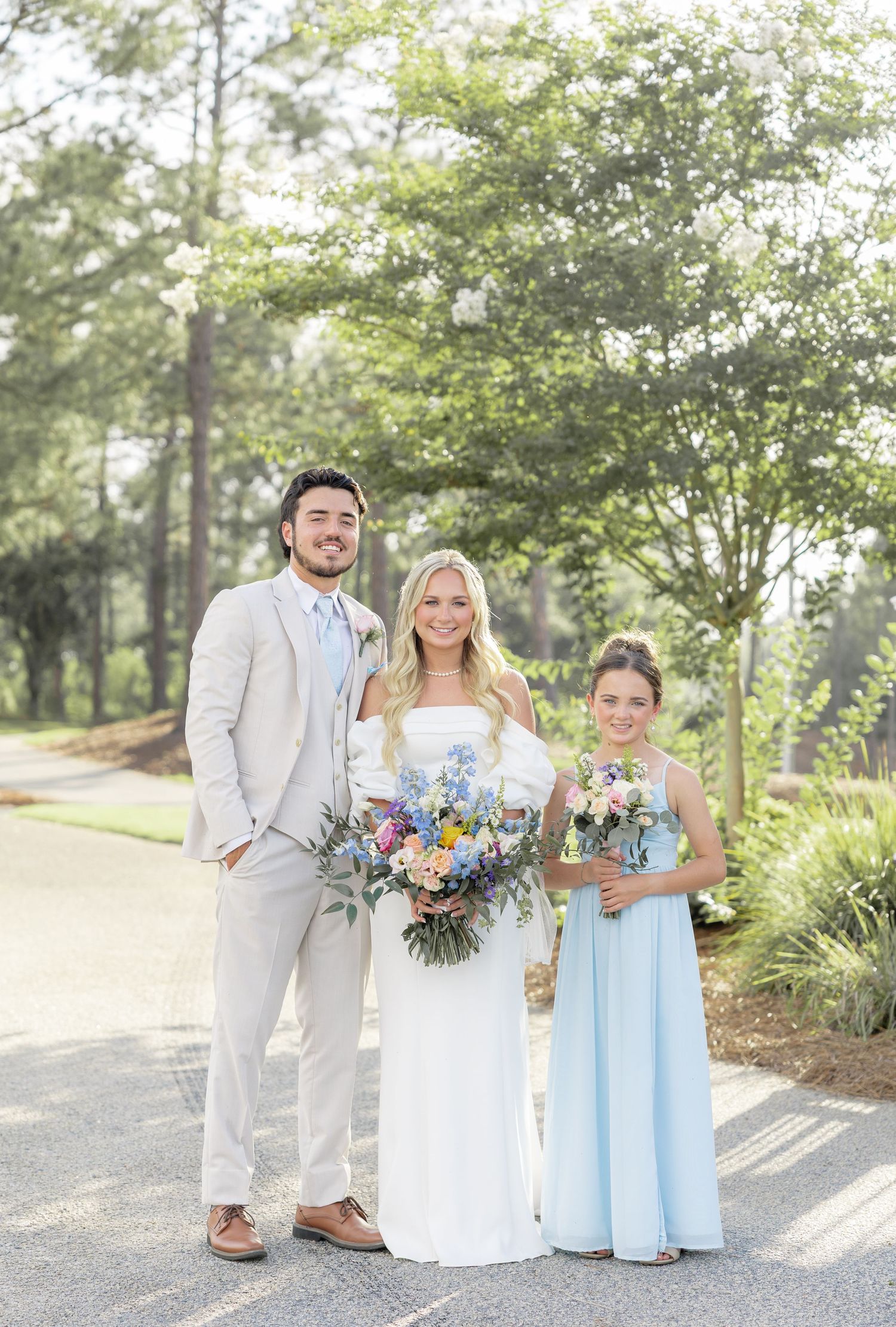 Groom in tan suit, blonde bride in white gown, and bridesmaid in blue holding bouquets outdoors.