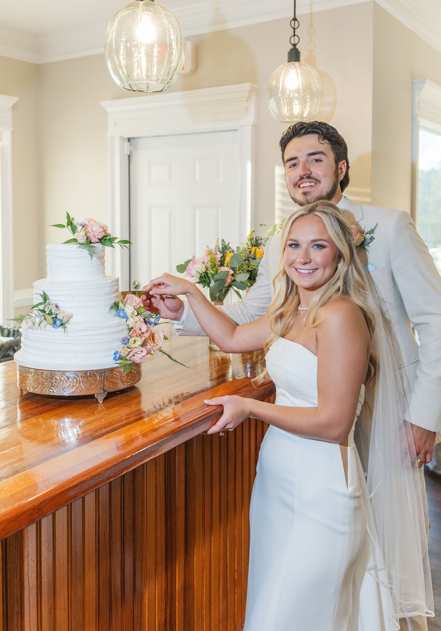 Smiling bride and groom cut white tiered wedding cake decorated with colorful flowers indoors.