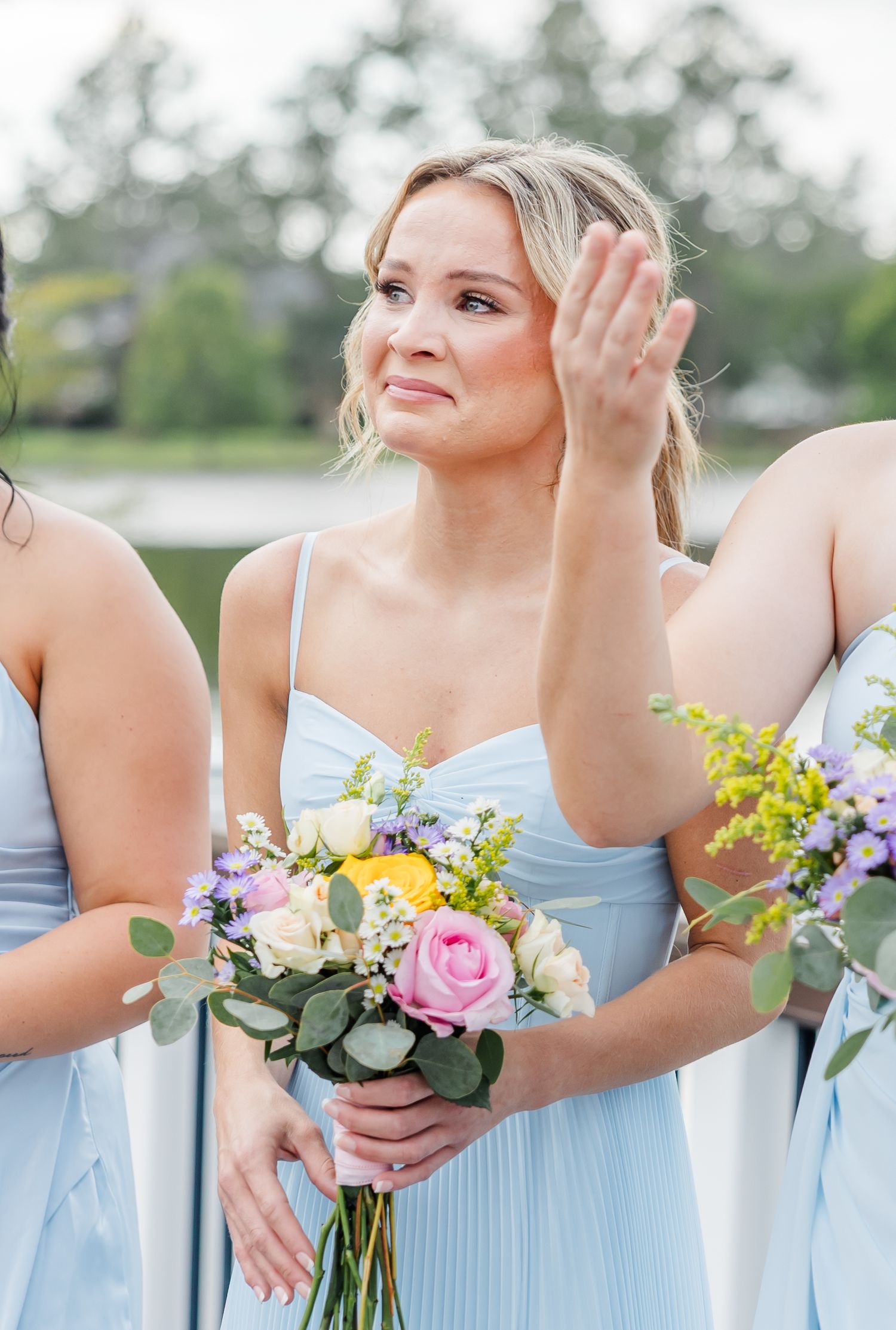Emotional bridesmaid in light blue dress holds floral bouquet during outdoor ceremony.