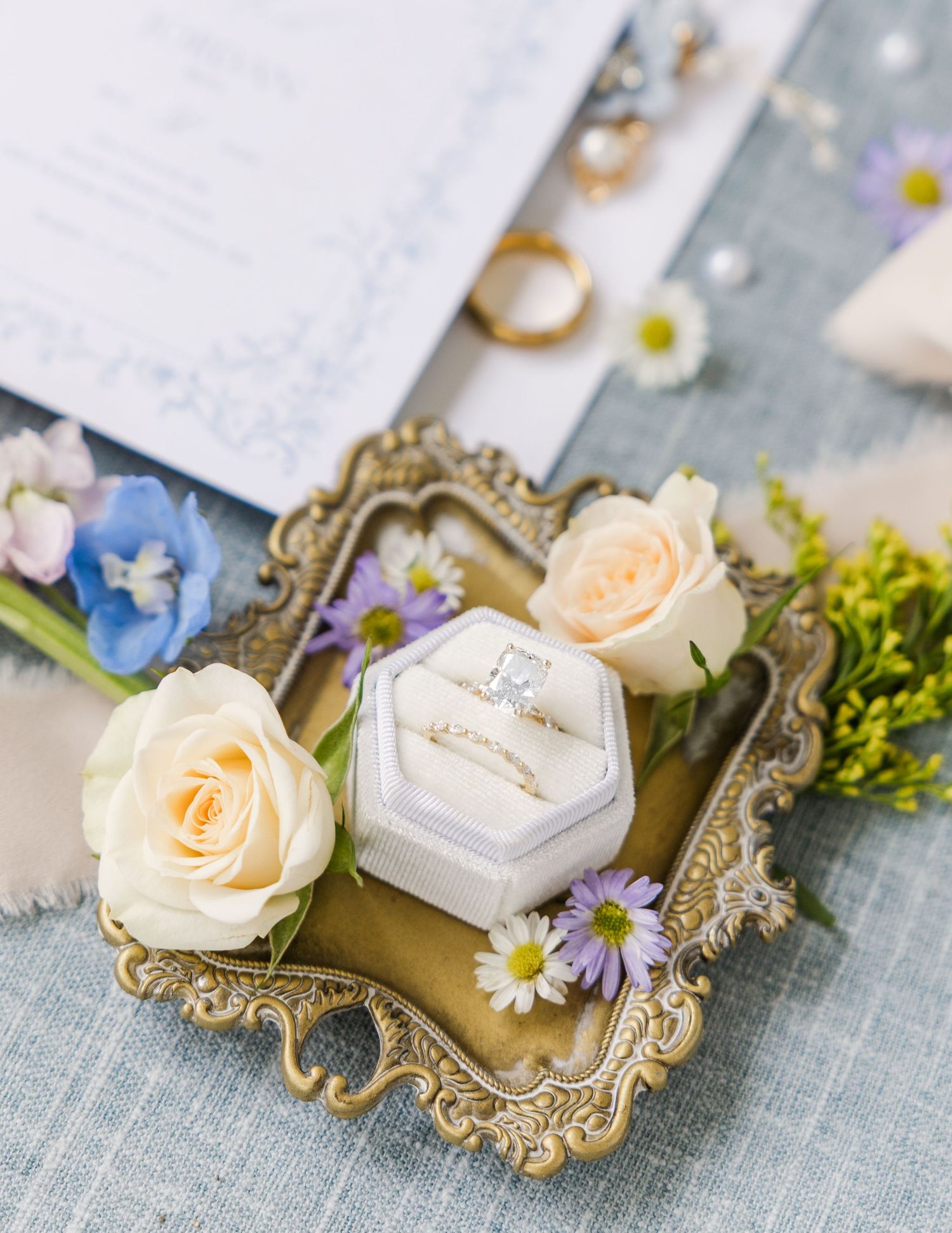 Gold ornate tray with wedding rings in velvet box, cream roses, and wildflowers on blue linen backdrop.