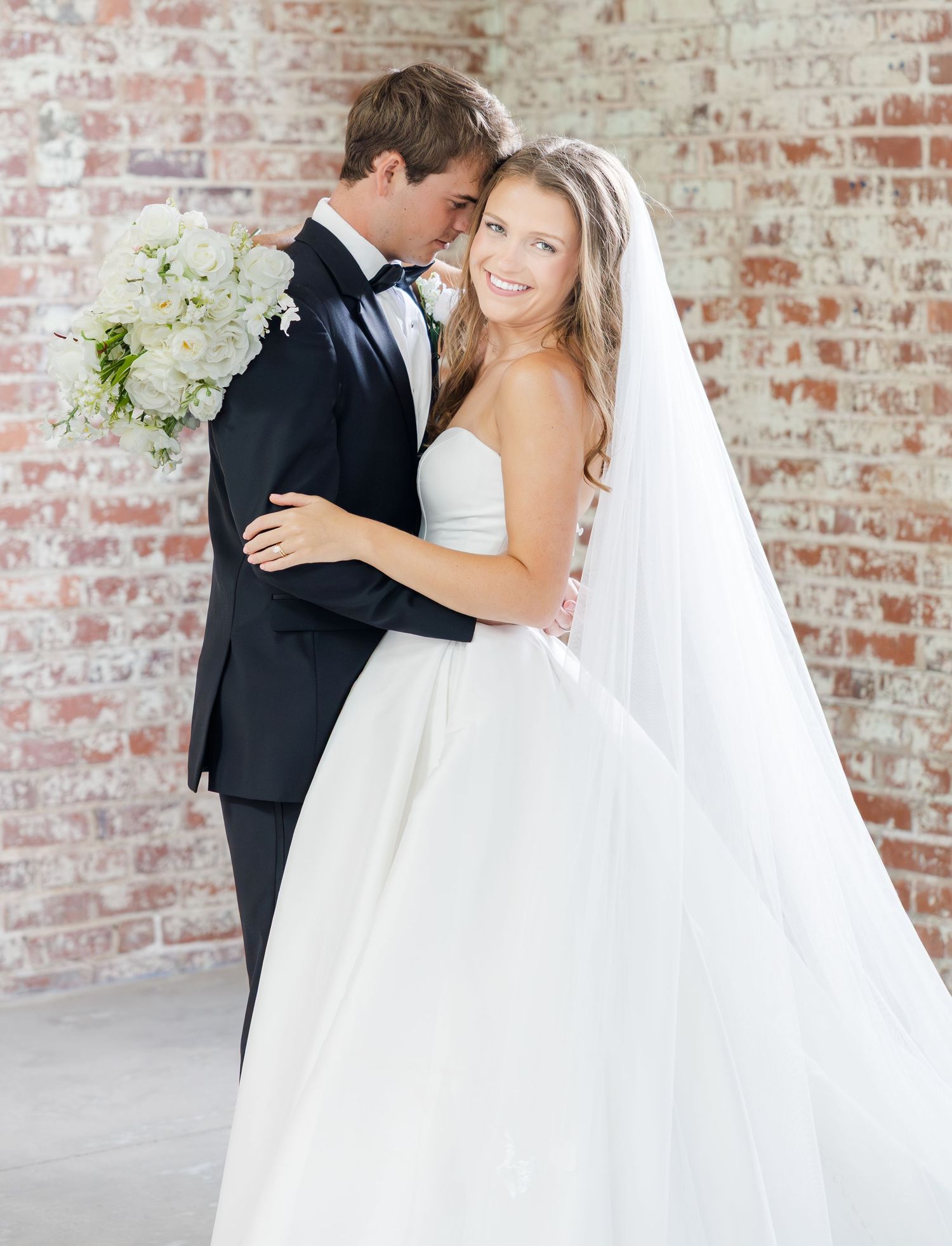Bride in strapless ballgown with long veil smiles as groom in black tuxedo embraces her against brick wall.