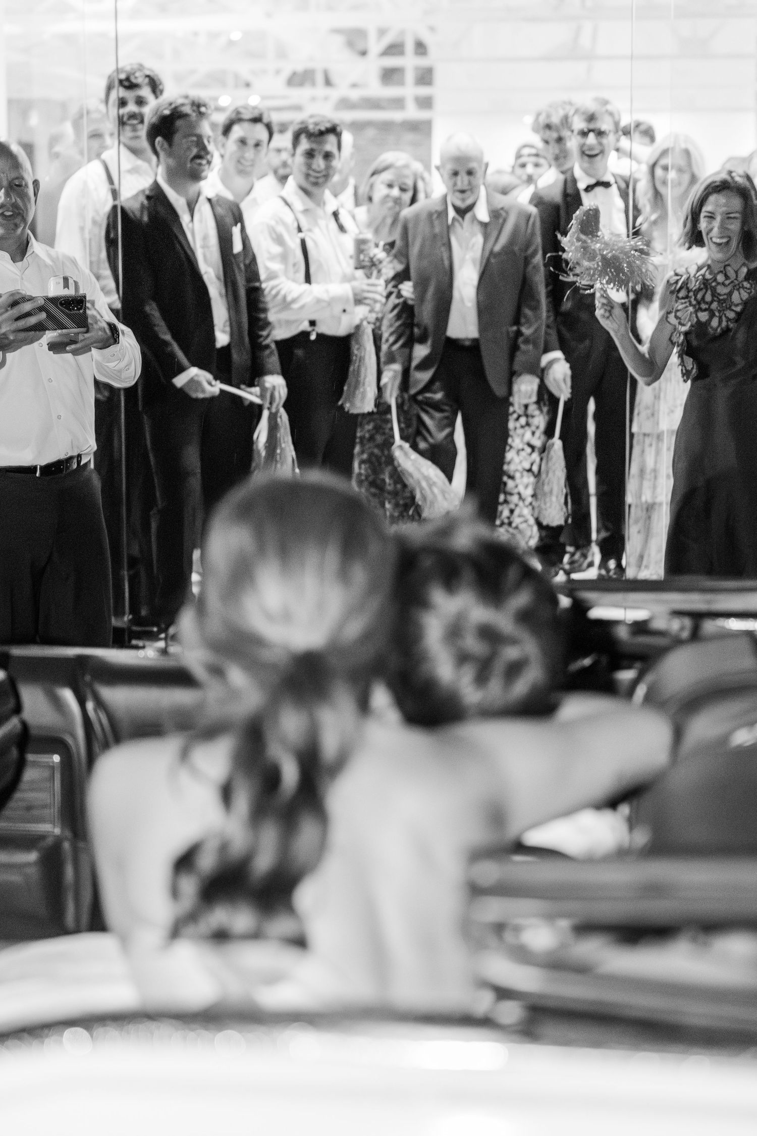 Black and white photo of bride and groom departing in convertible car as wedding guests cheer goodbye.