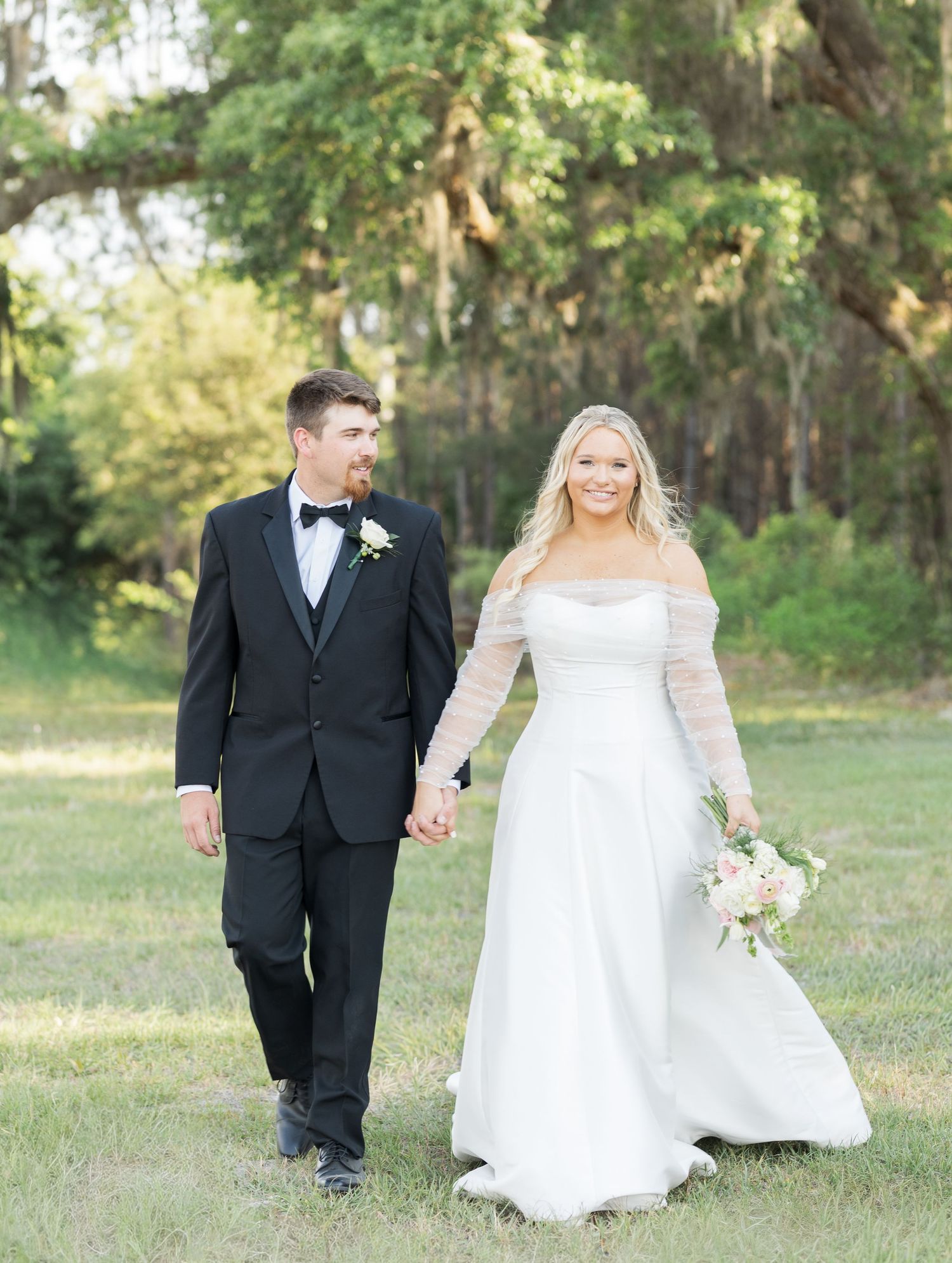 Bride and groom walking hand in hand outdoors, groom in black tuxedo, bride in off-shoulder gown.
