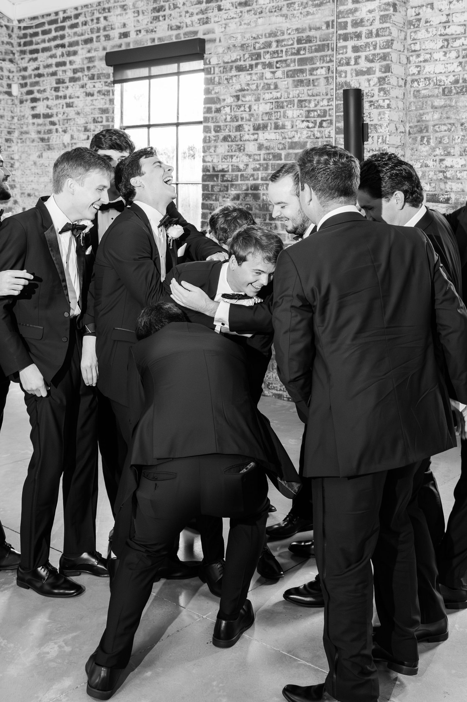 Groomsmen playfully pile on each other in a black and white candid wedding photo.