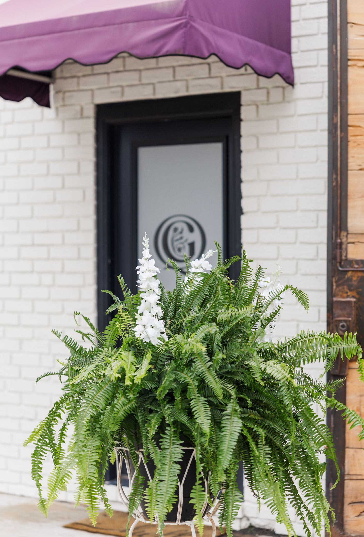 White brick building entrance with purple awning, monogram door sign, and lush fern planter.