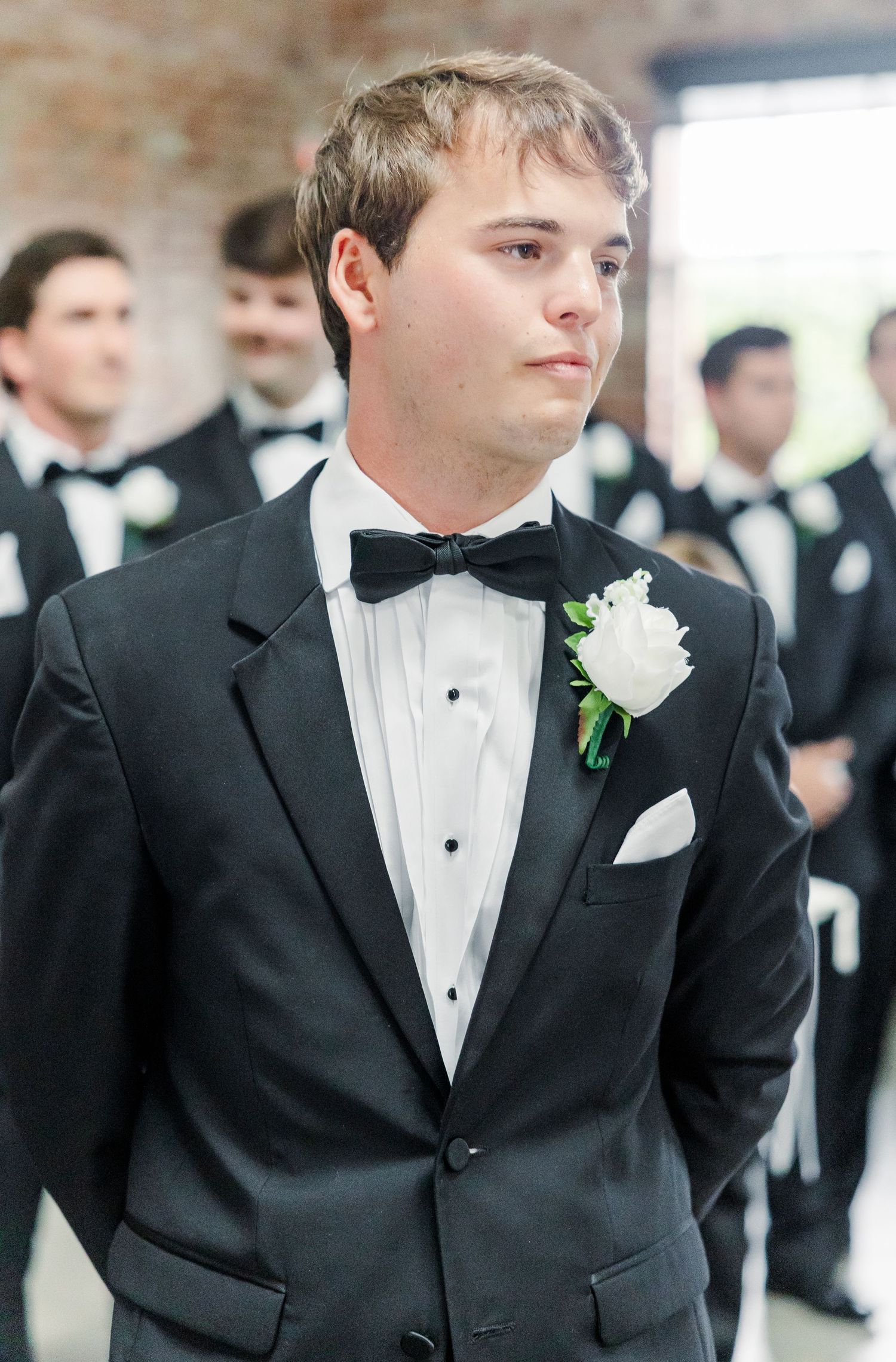Young groomsman in black tuxedo with white rose boutonniere stands at wedding ceremony.