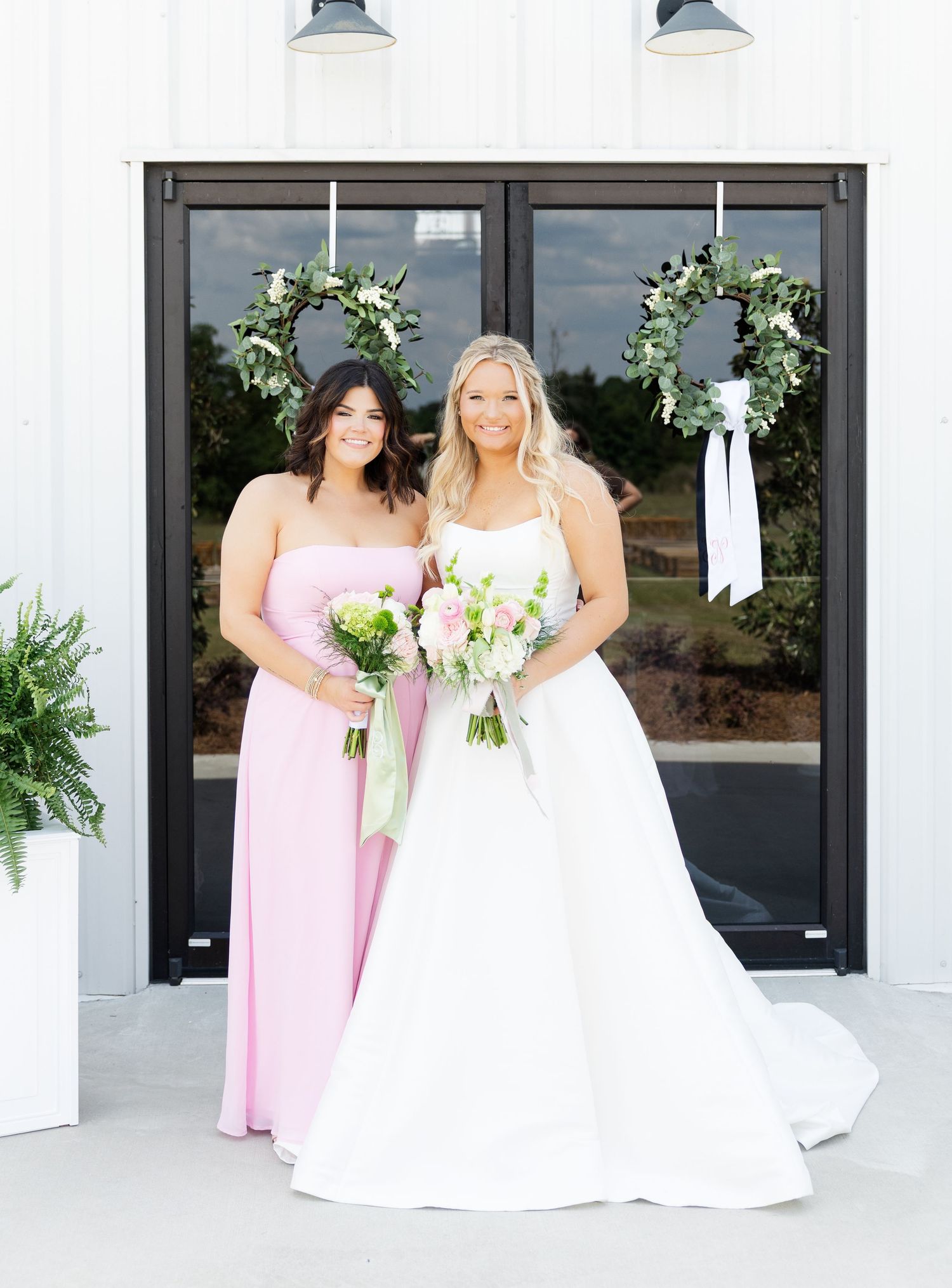 Bride in white gown and bridesmaid in pink dress holding bouquets outside white barn venue with floral wreaths.