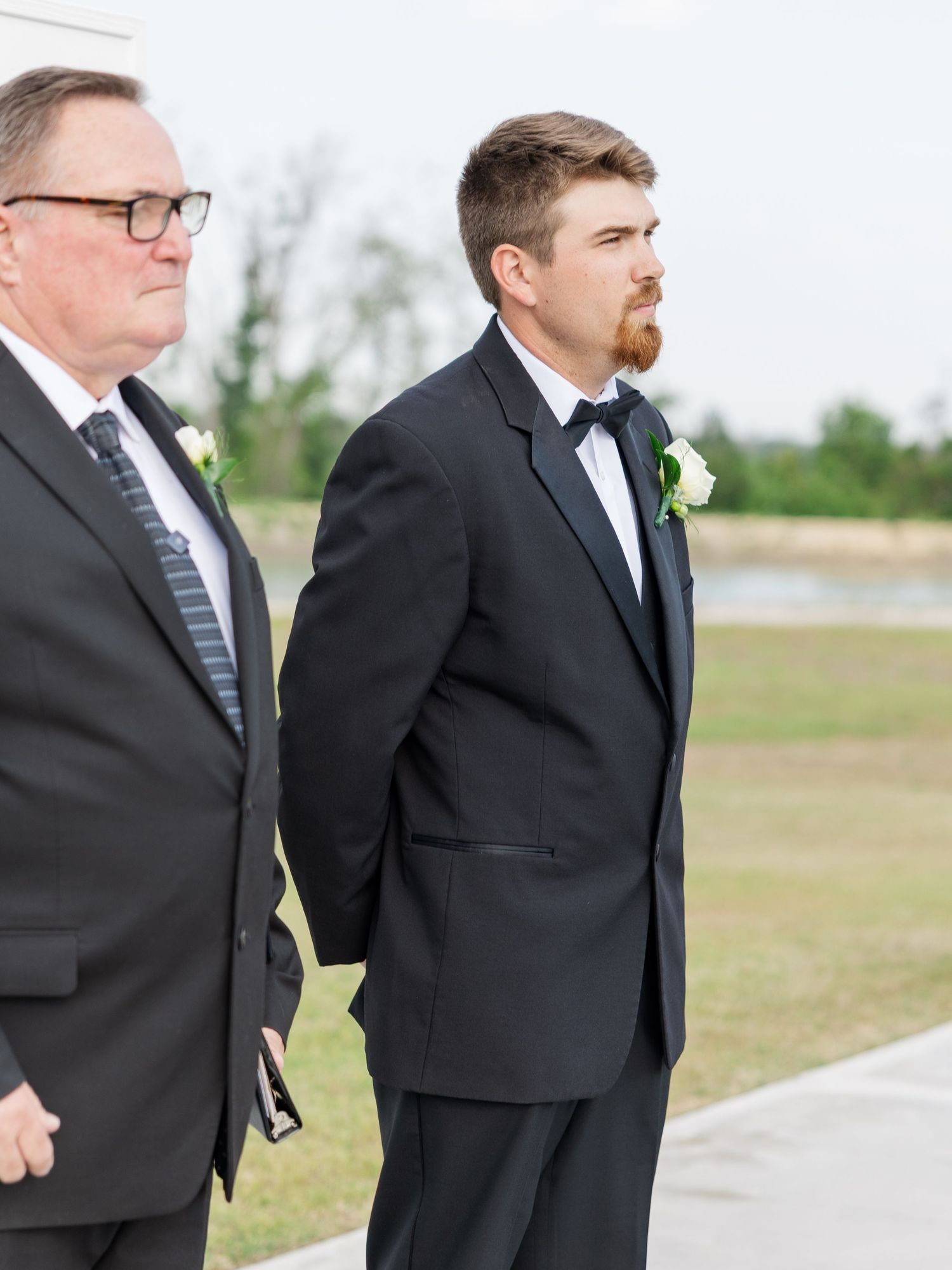 Groom in black tuxedo with boutonniere standing beside older man in suit at outdoor wedding ceremony.