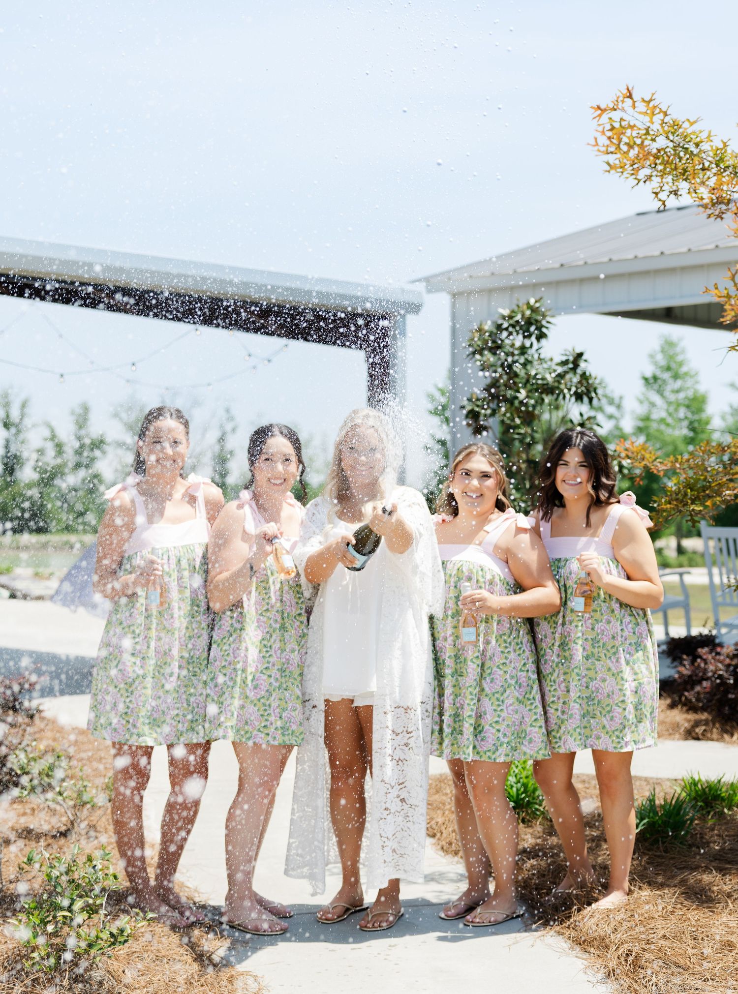 Bridesmaids in floral dresses celebrate with bride as champagne bottle pops outdoors.