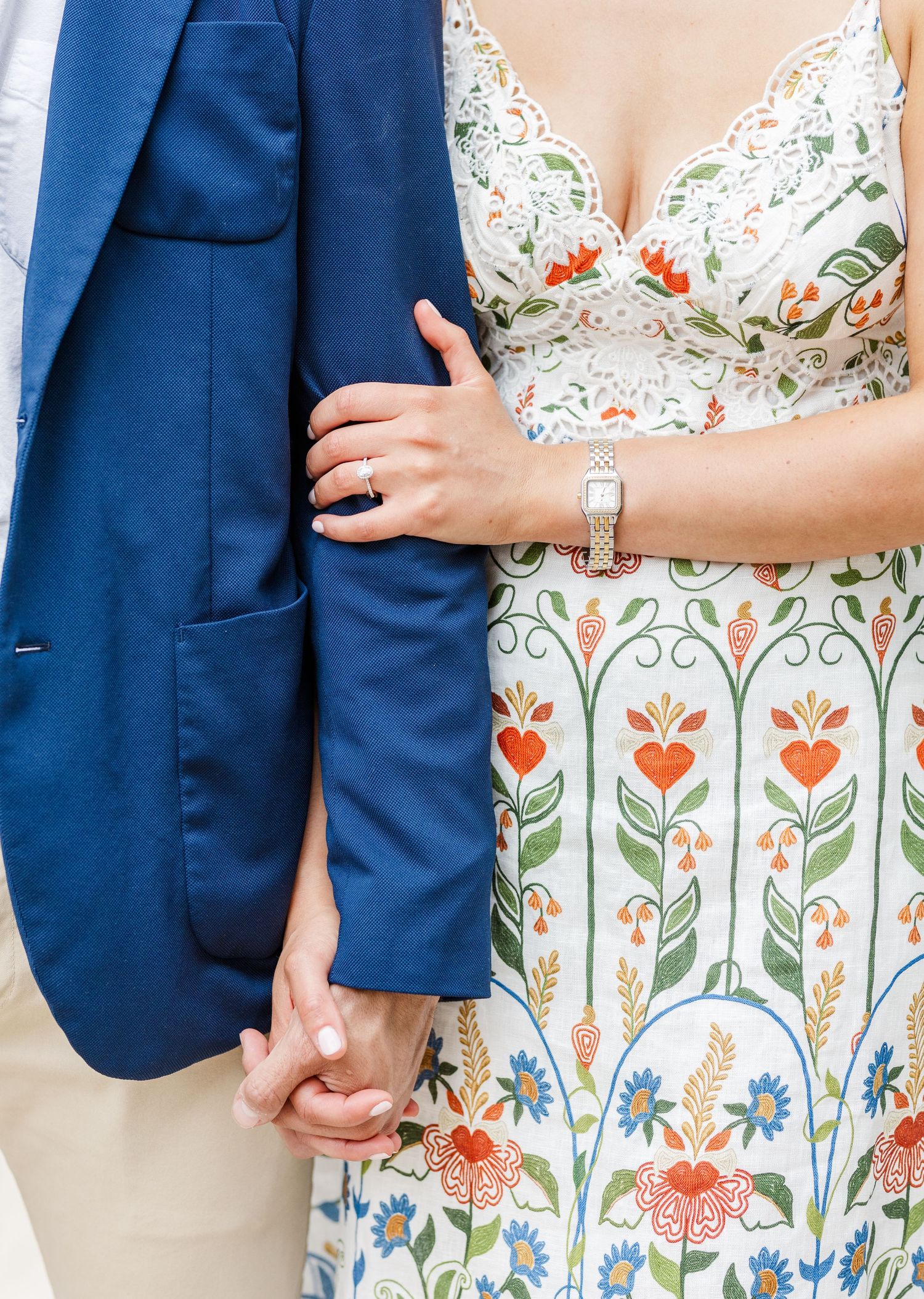Close-up of couple holding hands, woman wearing floral dress and engagement ring, man in navy blue blazer.