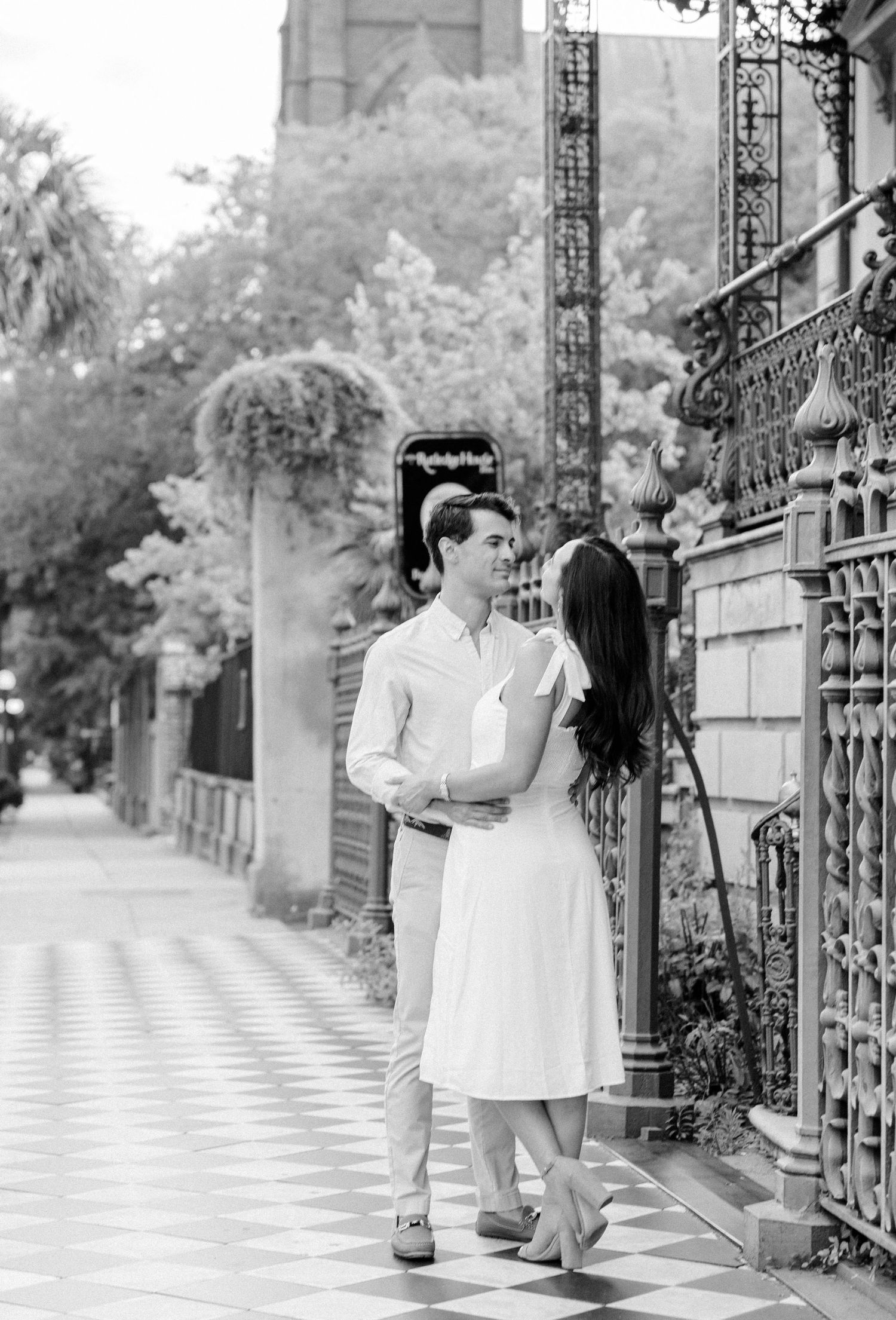Black and white photo of a couple embracing on a charming sidewalk beside ornate iron gates and lush greenery.