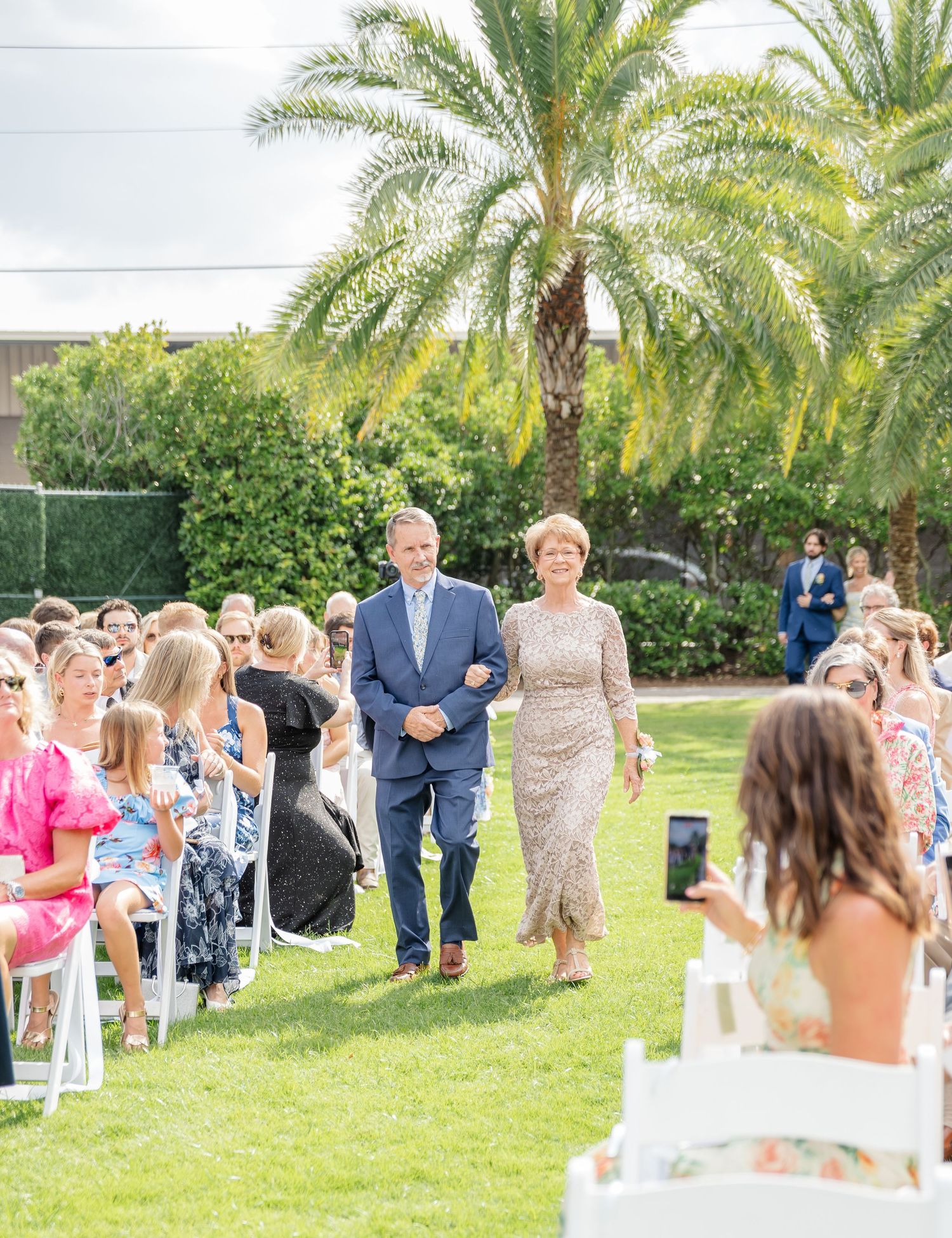 Outdoor wedding ceremony setup with white chairs and palm trees in a lush garden setting.