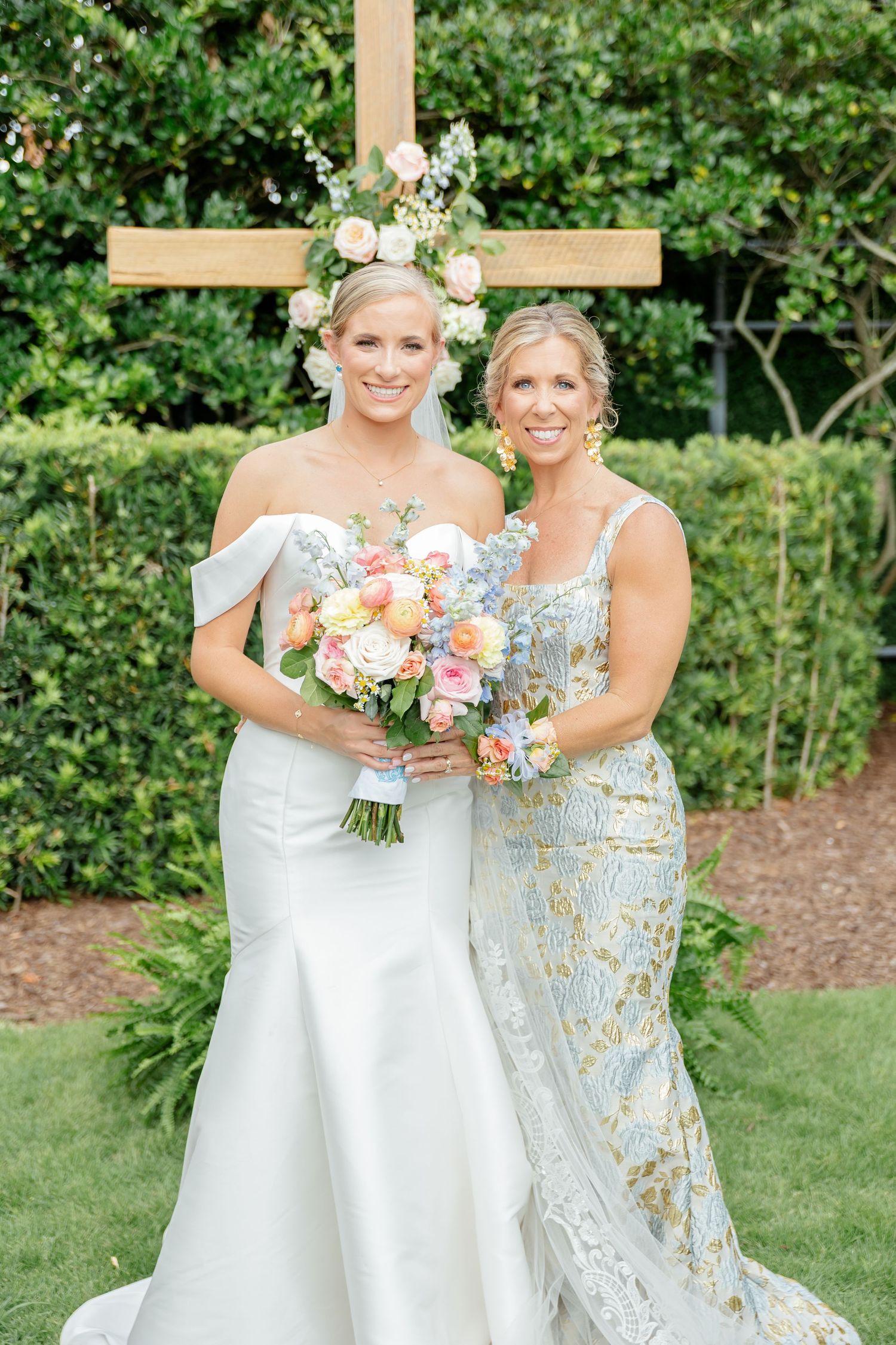 Elegant wedding photo outdoors with bride in white gown and bridesmaid in blue floral dress holding pastel bouquets.