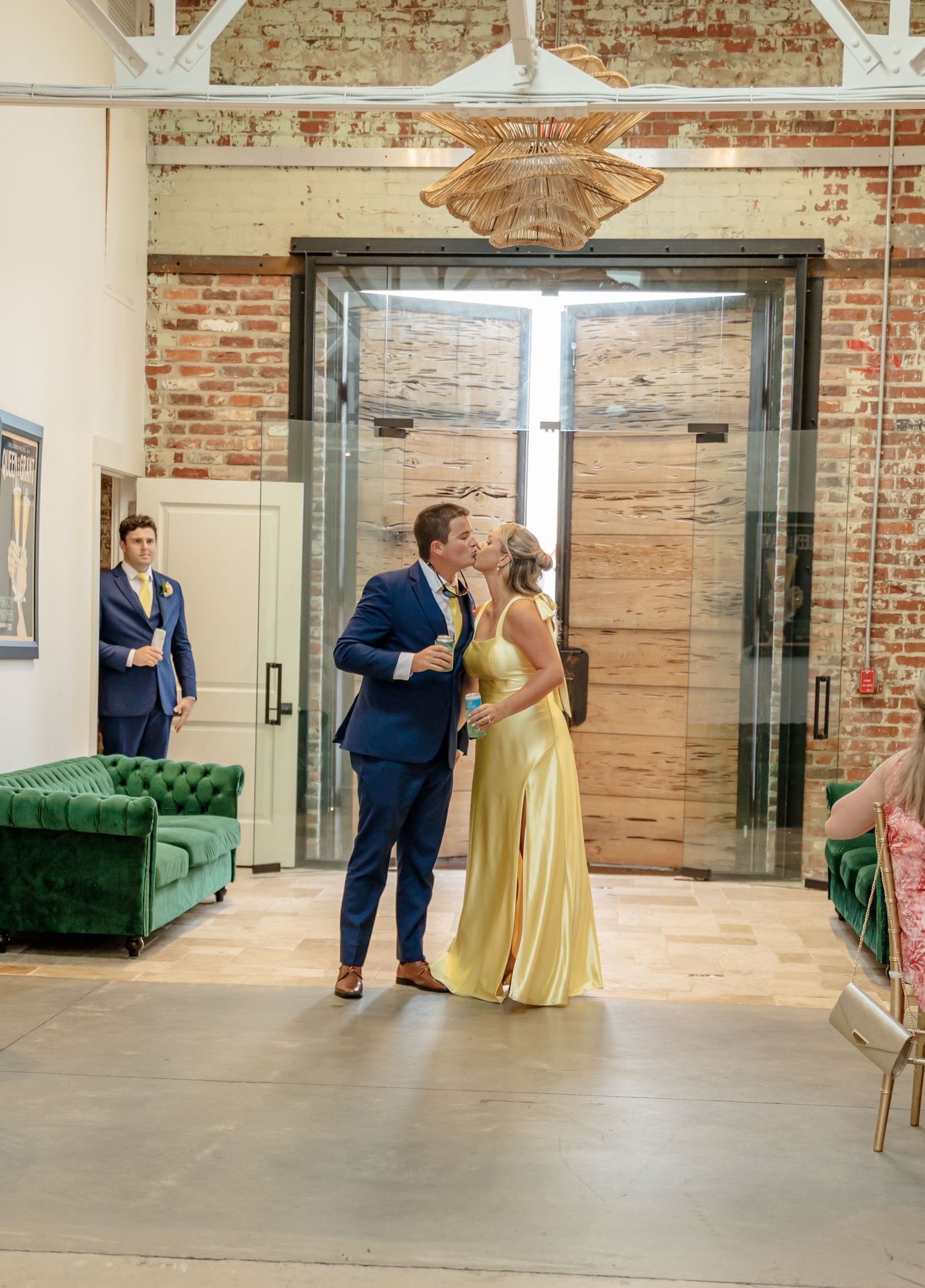 A couple in formal attire, with a yellow dress and navy suit, share a dance moment in an industrial brick venue space.