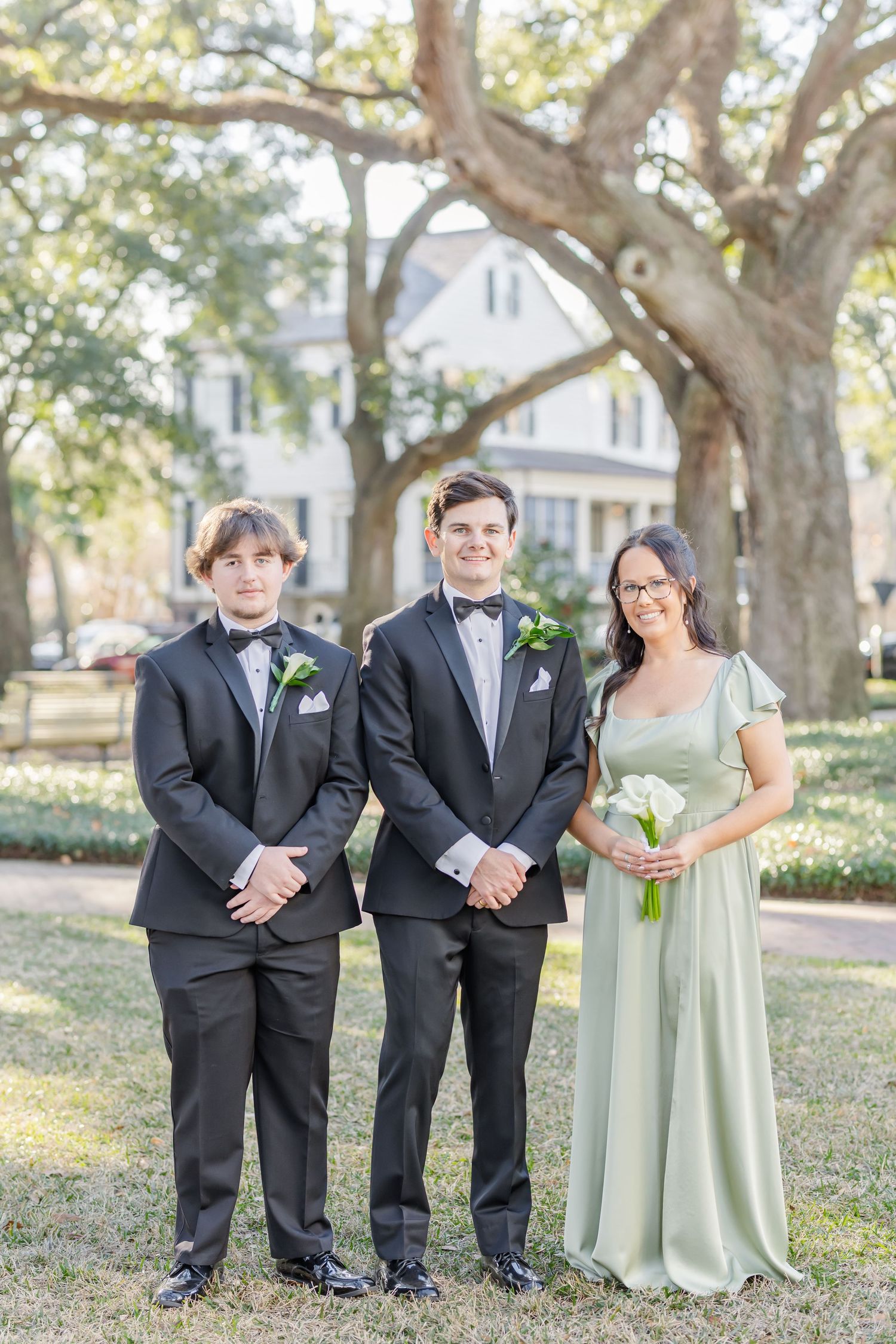Wedding party members in black tuxedos and sage green dresses pose under trees.