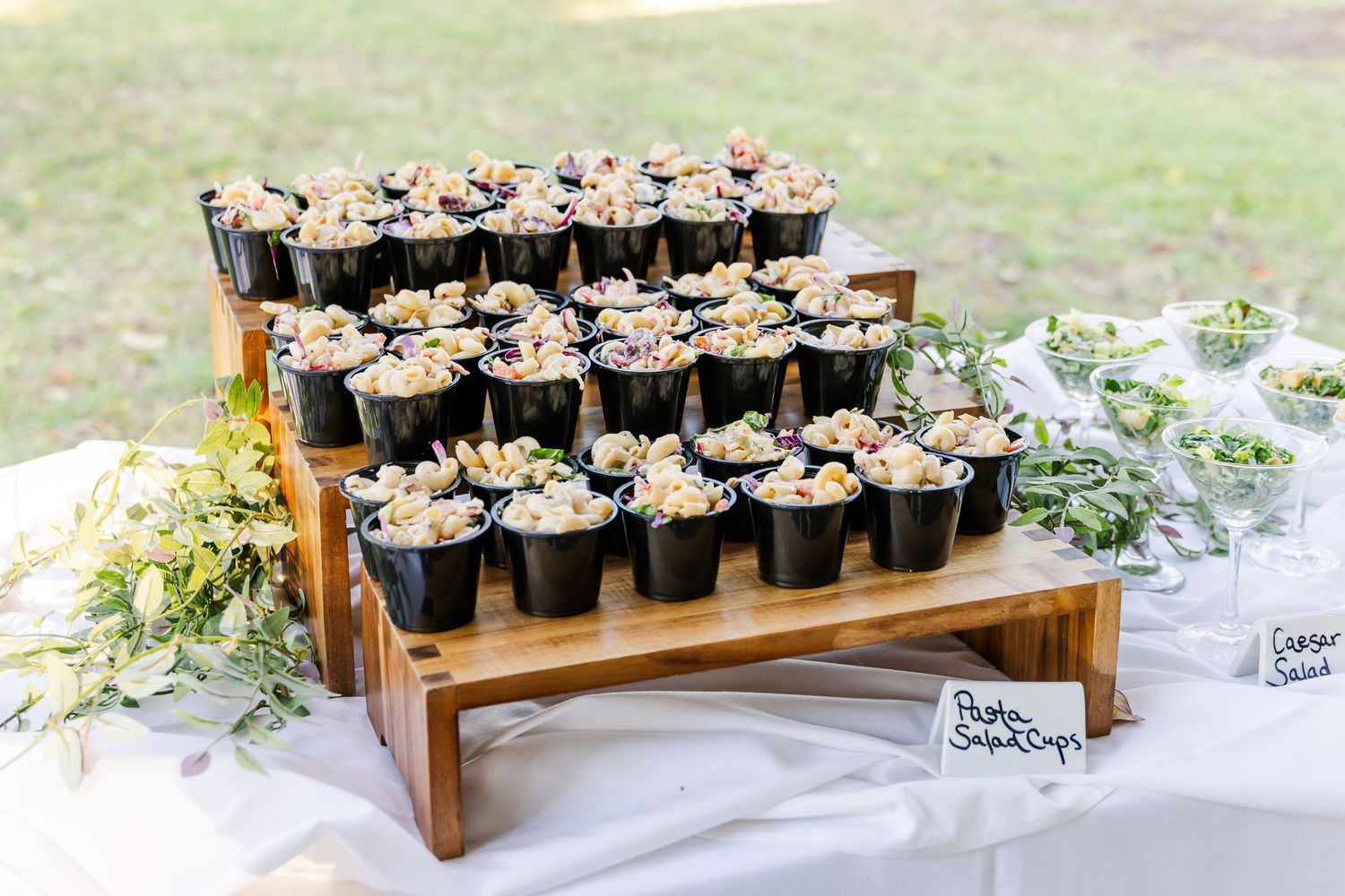 Rustic wooden display stand holding rows of black potted favors with green foliage accents.