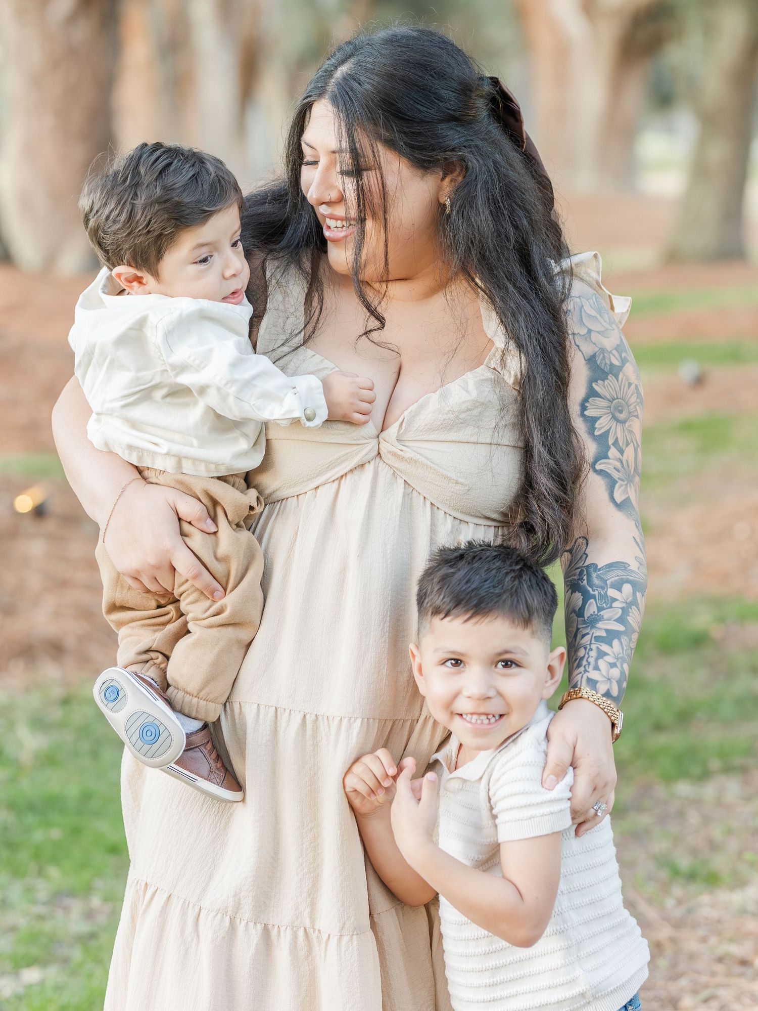 A mother in a flowing cream dress shares tender moments with her children in a wooded setting.