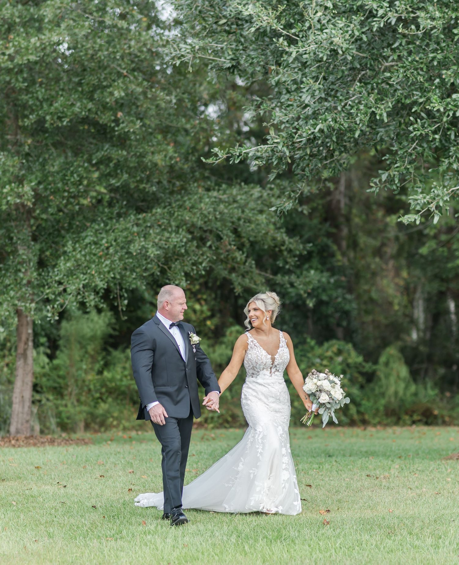 A wedding couple shares playful moments on a lush green lawn surrounded by trees.