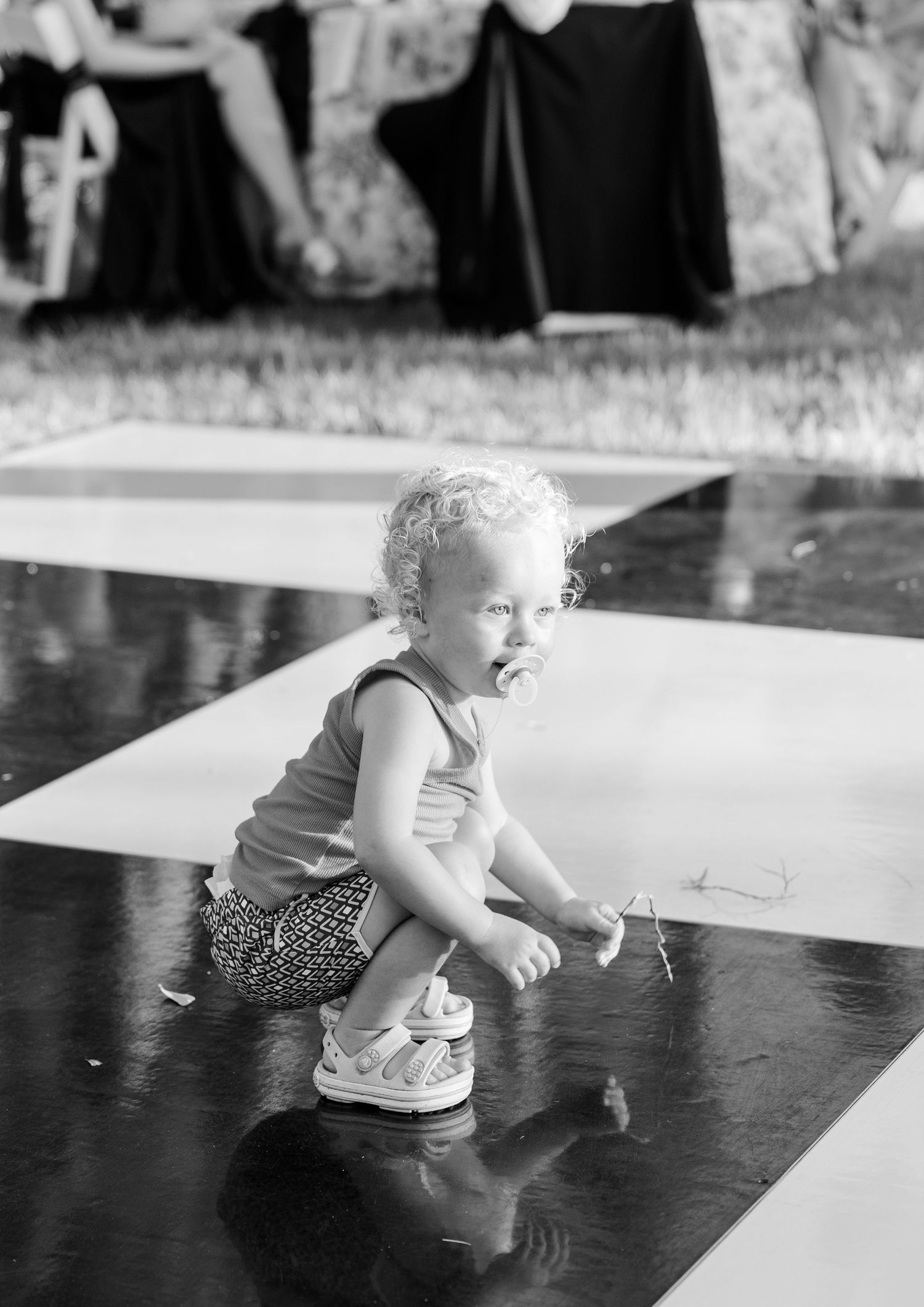 A toddler with curly hair crouches on a dance floor at an outdoor wedding reception in black and white photo.