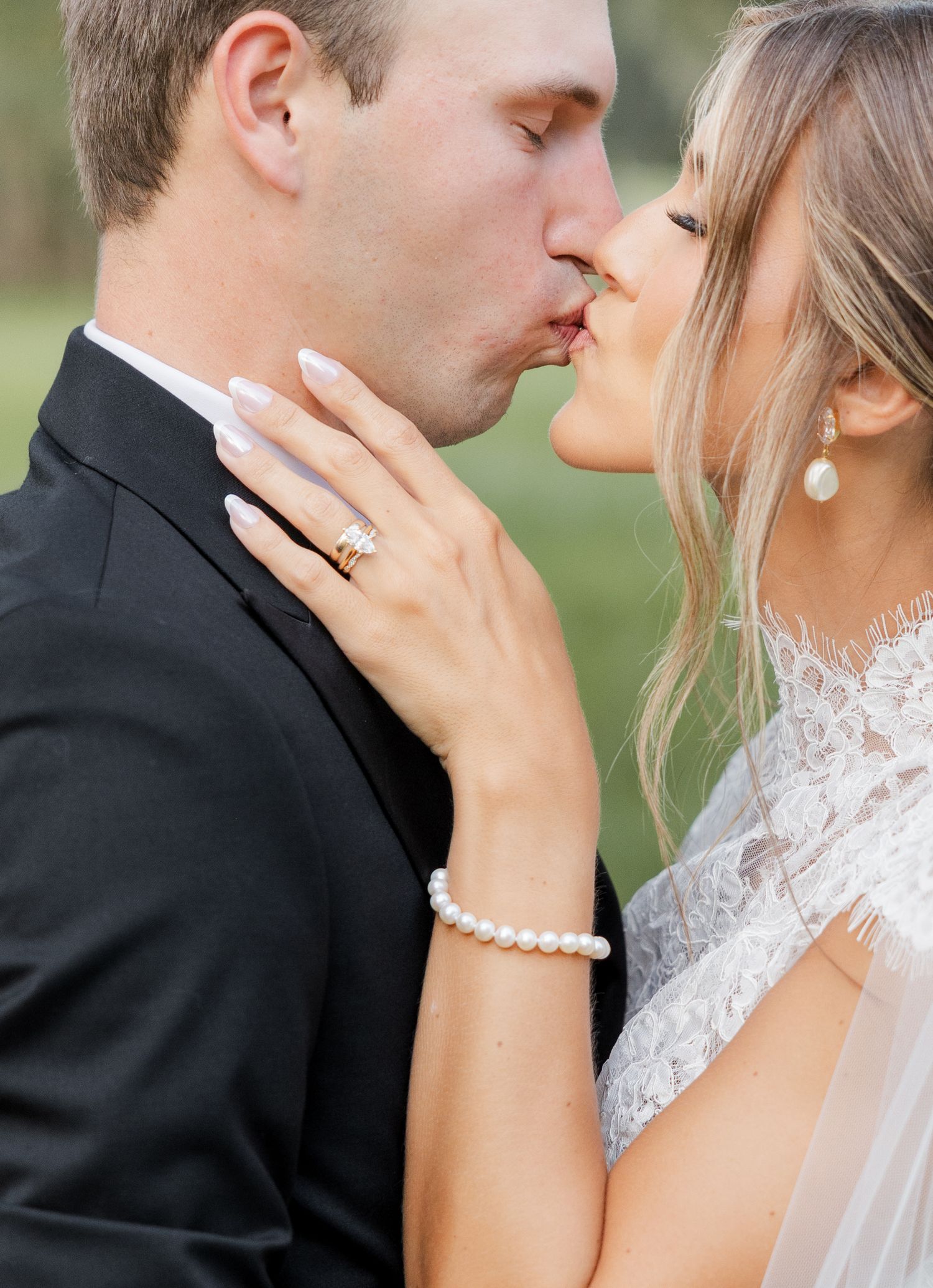 A bride and groom share a romantic kiss, she wears a pearl bracelet, ring, and lace gown on their wedding day.