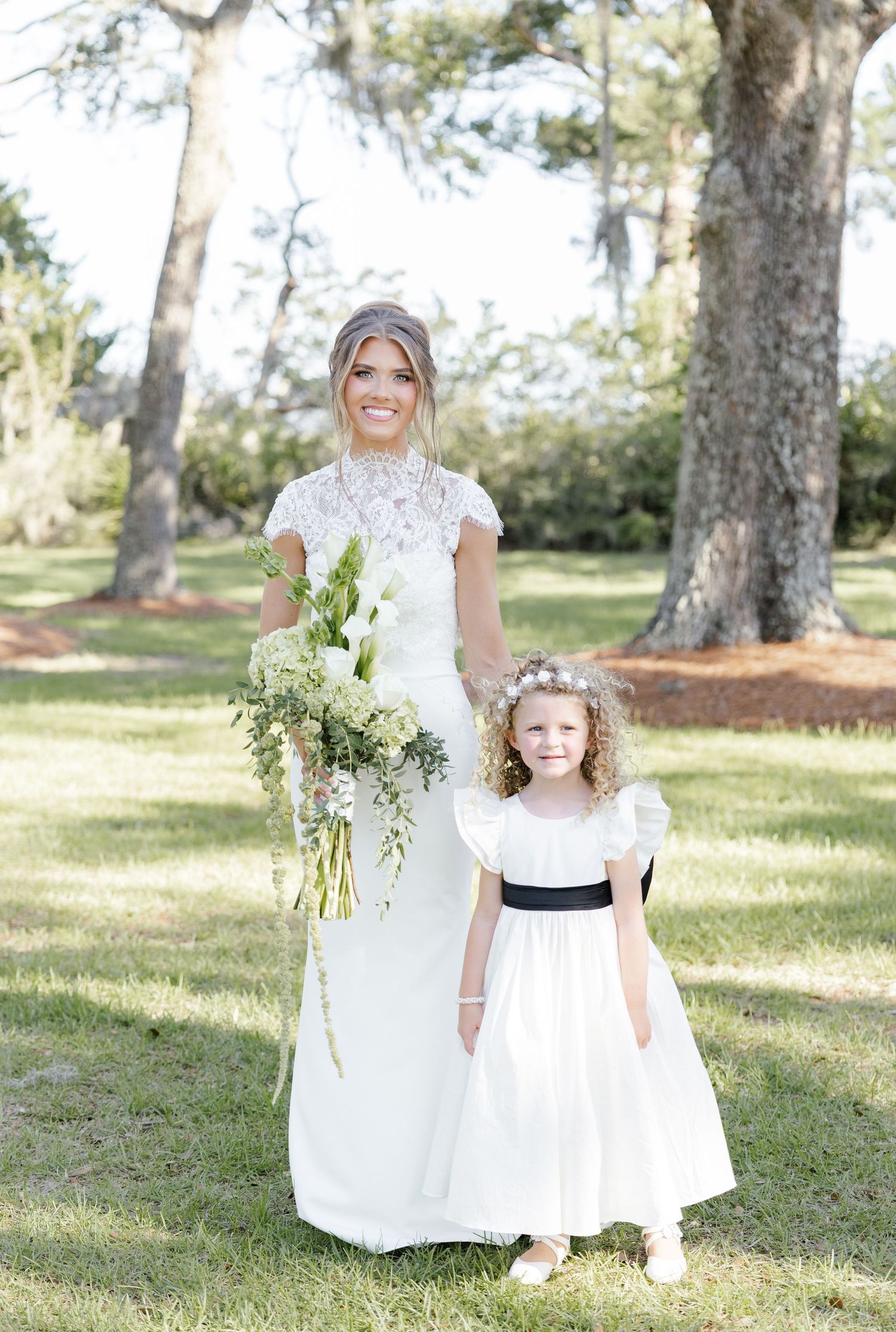 Bride in lace cap sleeve wedding gown poses with flower girl in white dress with black sash on green lawn.
