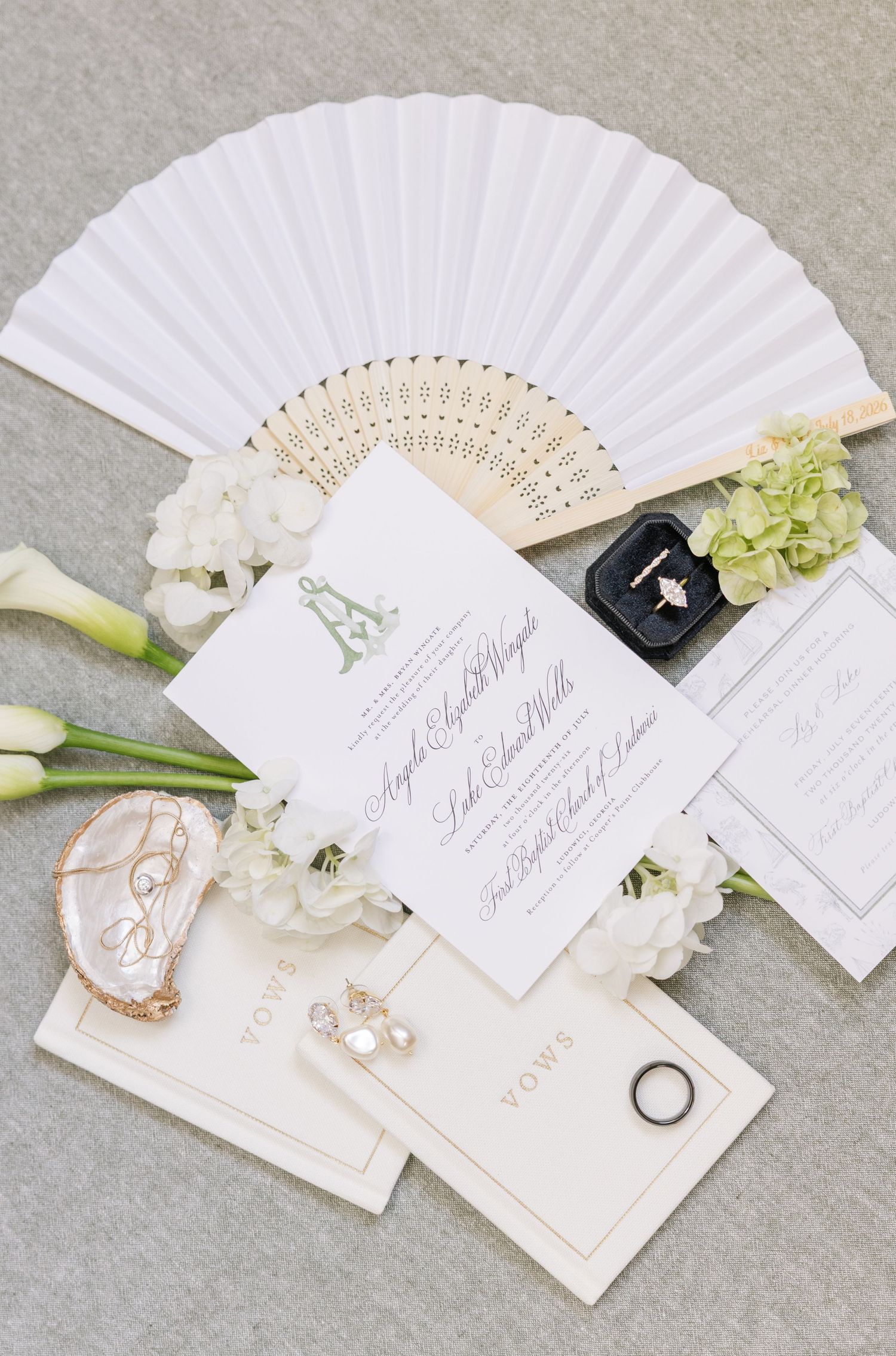 Elegant wedding flat lay with white fan, floral invitations, vow cards, pearl earrings, and rings on a neutral background.