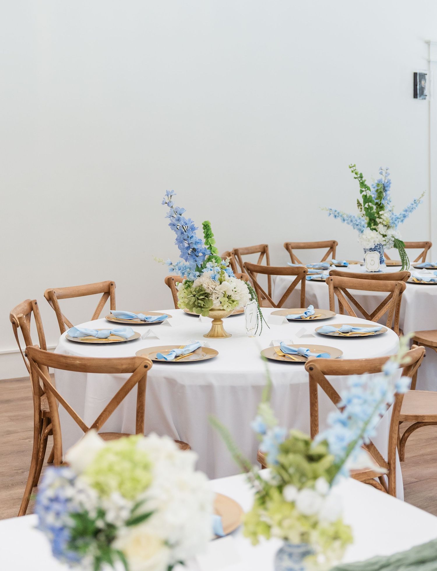 Wedding reception tables with white linens, gold chargers, blue napkins, and blue floral centerpieces.