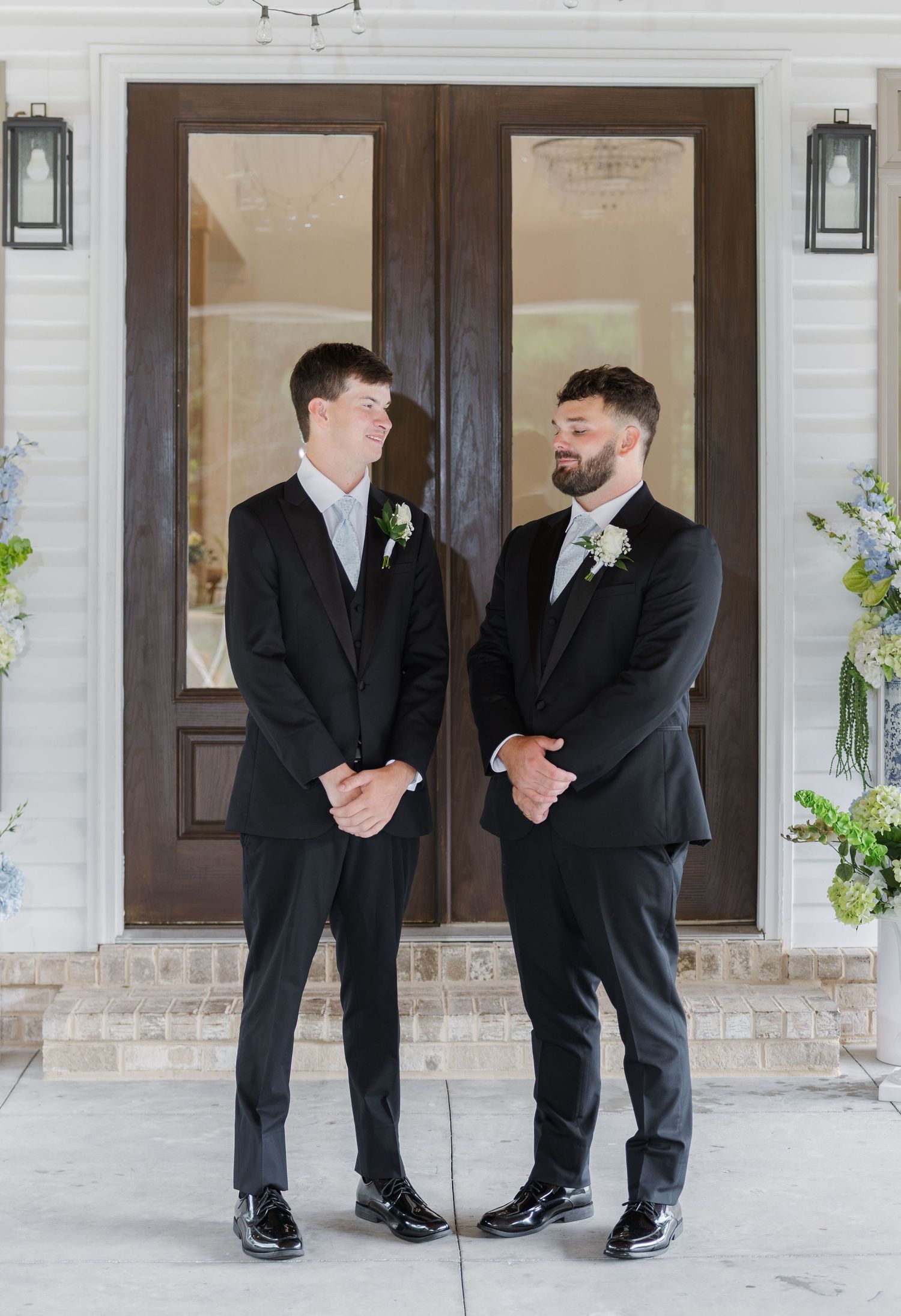 Groom and groomsman in black suits with light blue ties outside wooden double doors.