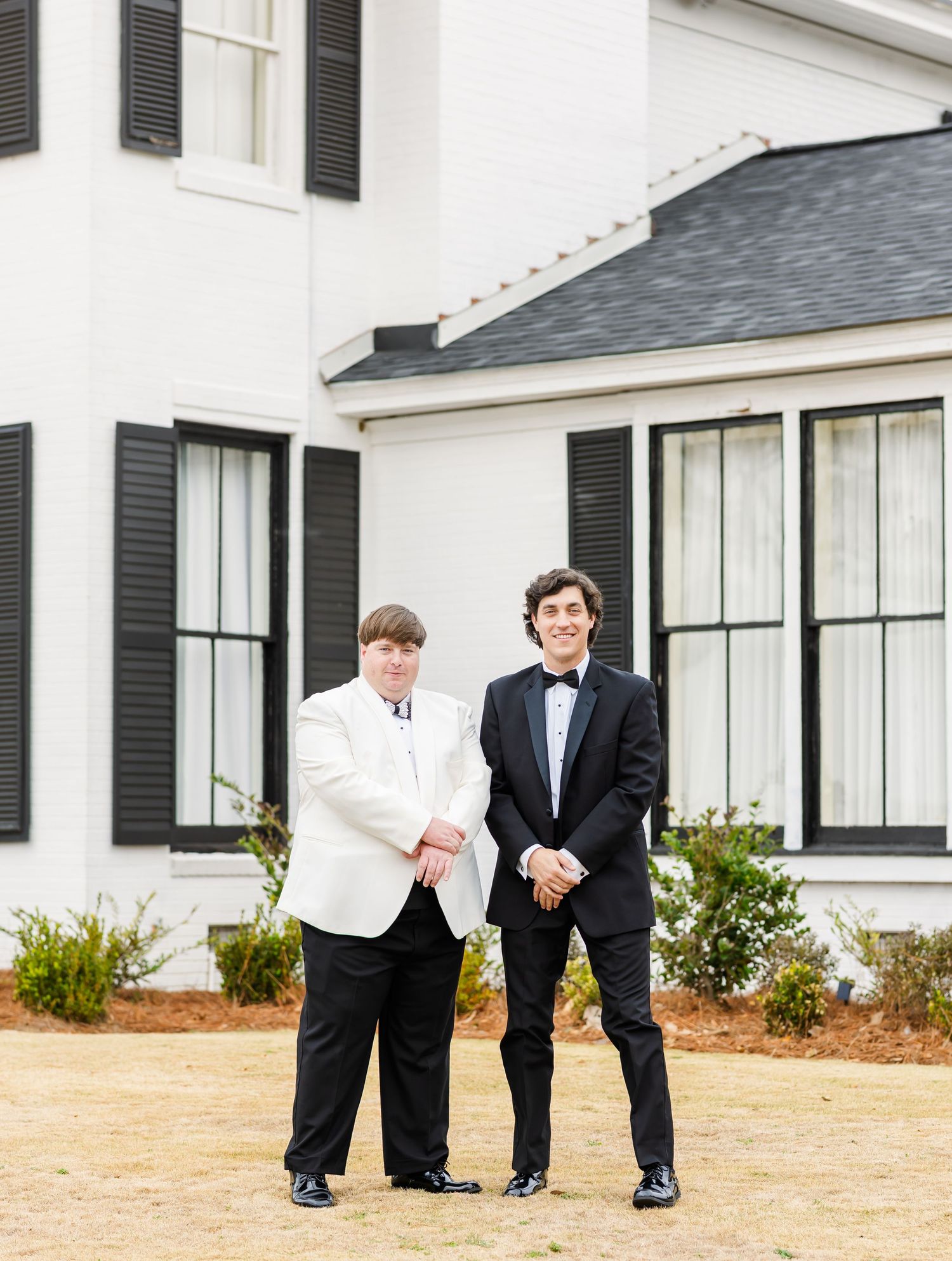 Groom in white jacket and groomsman in black tuxedo pose together outside white Southern wedding venue.