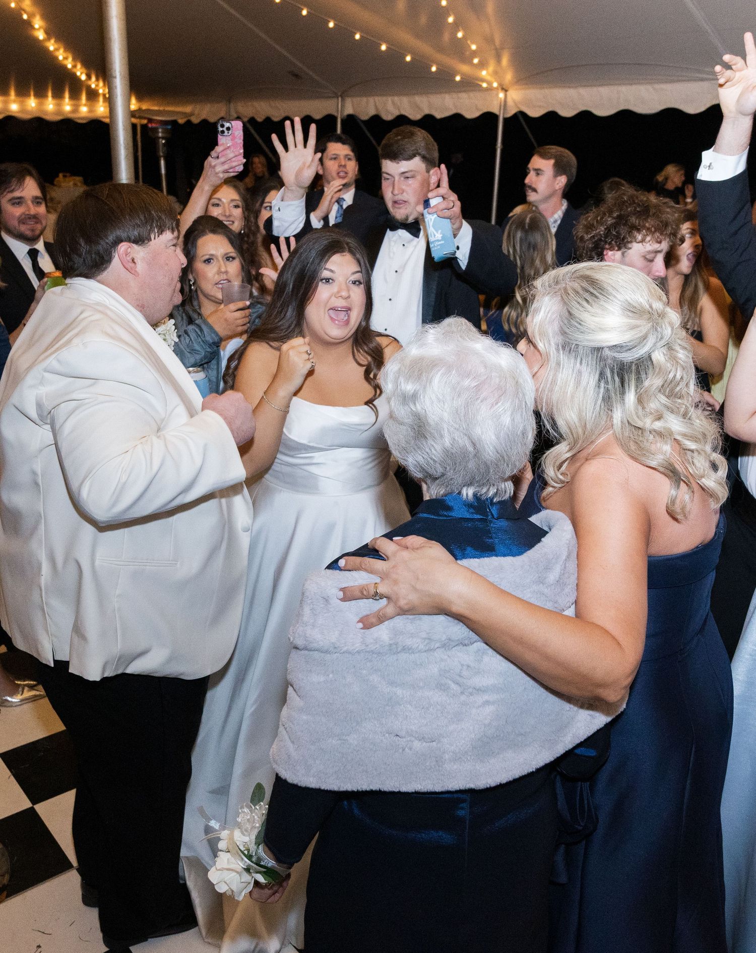 Joyful bride dancing with family and friends under string lights at lively wedding reception.