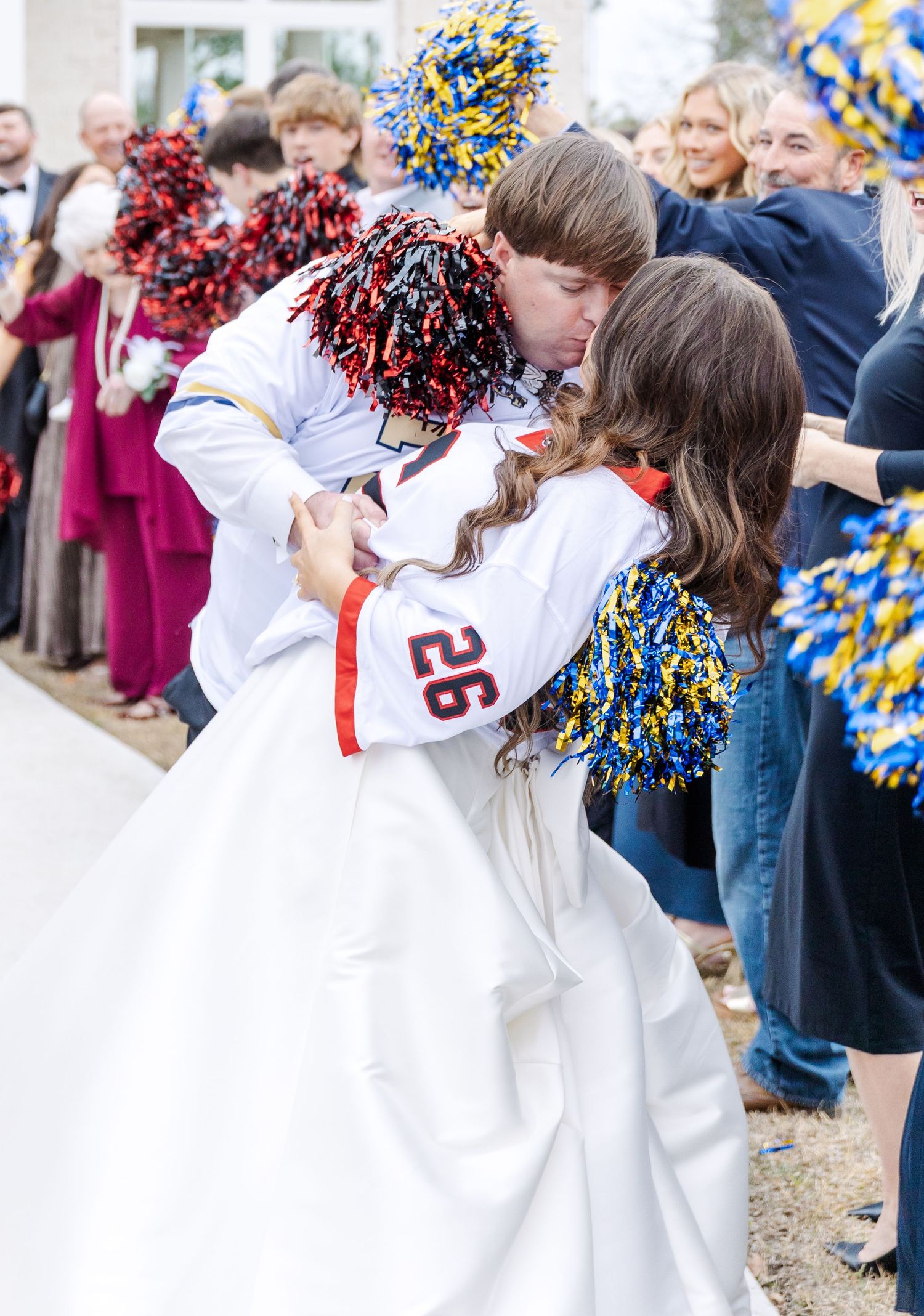 Groom in football jersey number 26 dips bride in wedding dress while guests wave colorful pom-poms outside chapel.