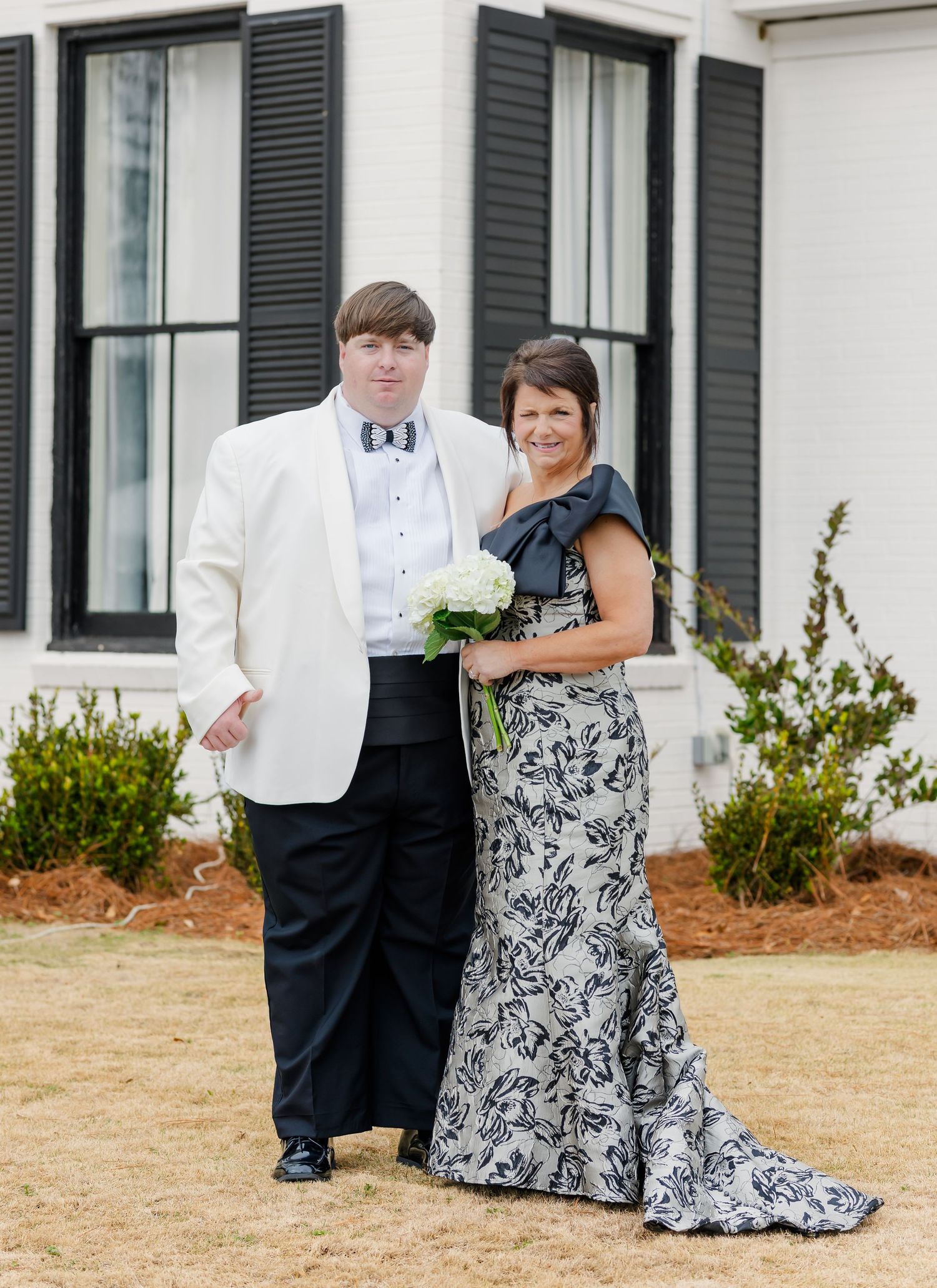 Young man in white tuxedo jacket poses with woman in black floral gown outside white building.