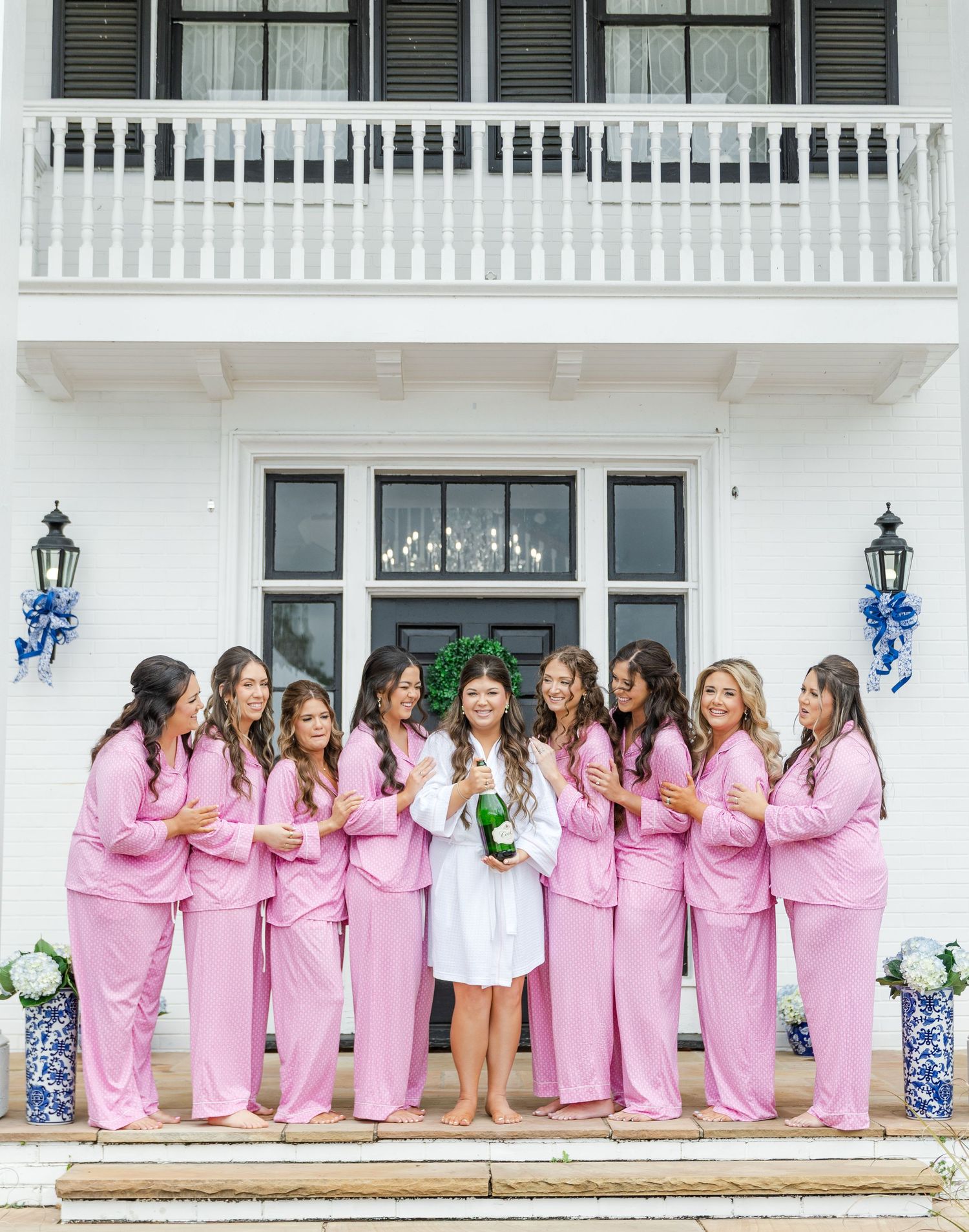 Bridesmaids in pink pajamas celebrate with bride in white robe holding champagne bottle outside white mansion.