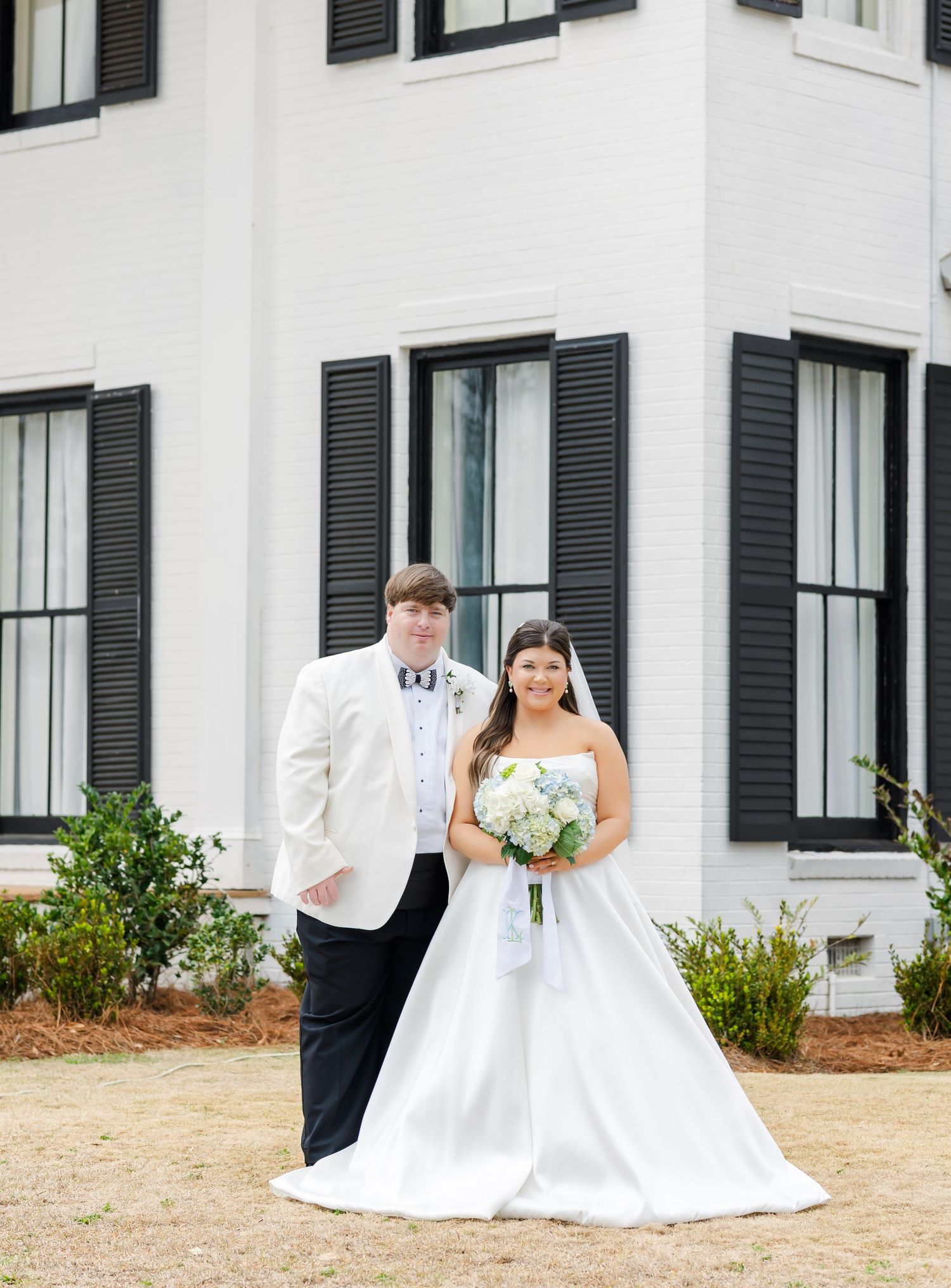 Bride and groom posing together outside a white Southern mansion with black shutters on their wedding day.