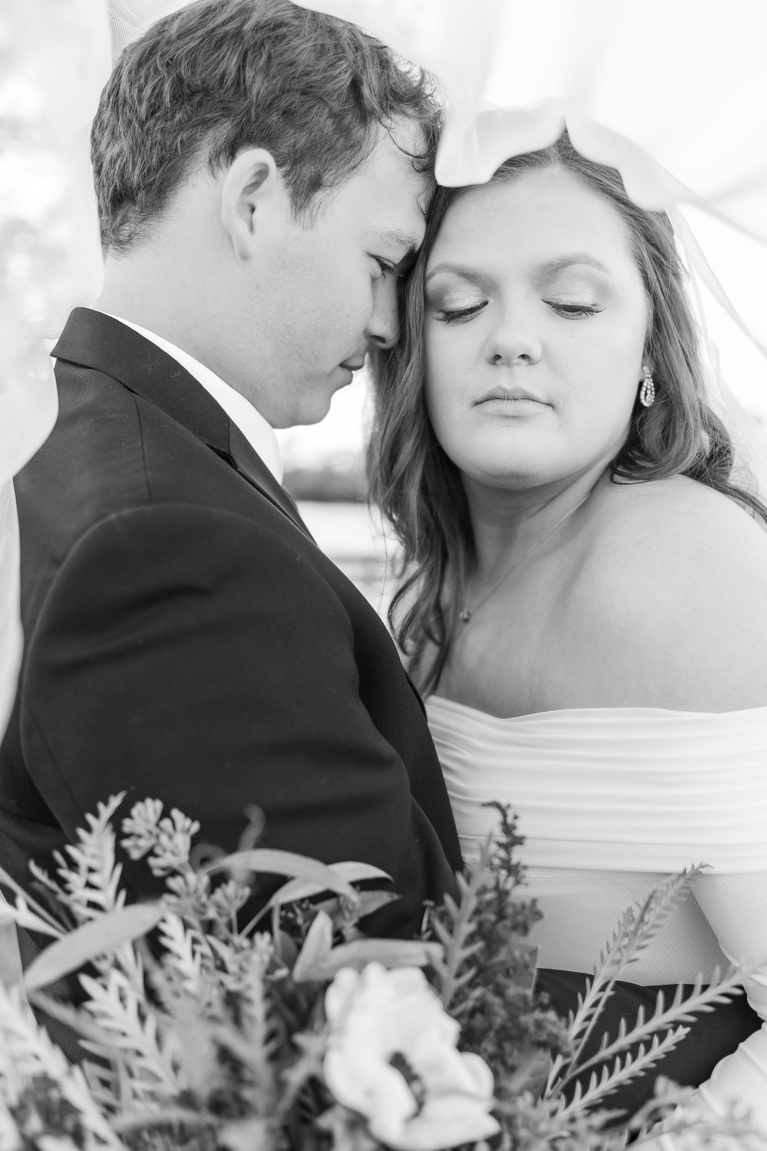 An intimate black and white wedding portrait showing a tender moment between newlyweds with a bouquet.