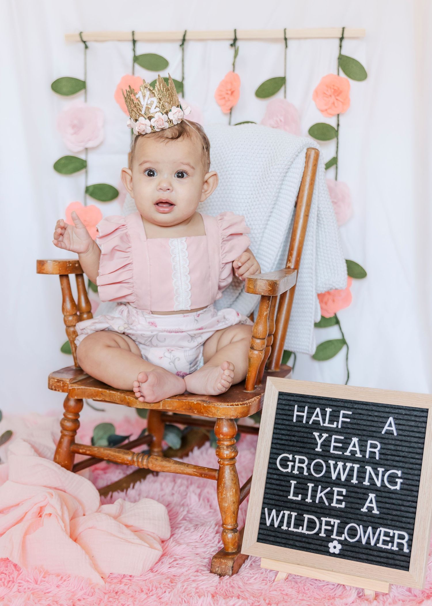 Six month milestone photo with baby in rocking chair next to letterboard sign and floral backdrop.