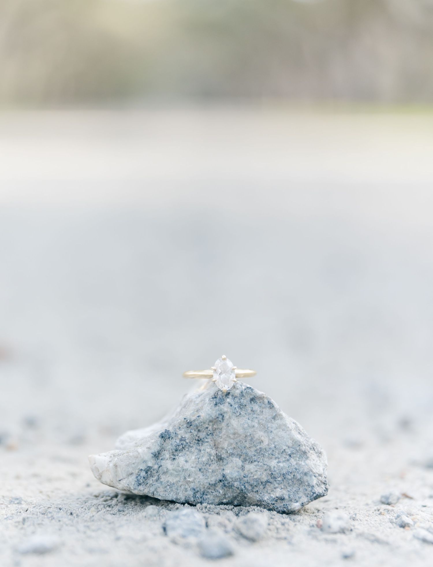 A diamond engagement ring sits atop a light-colored stone in dreamy, soft focus.
