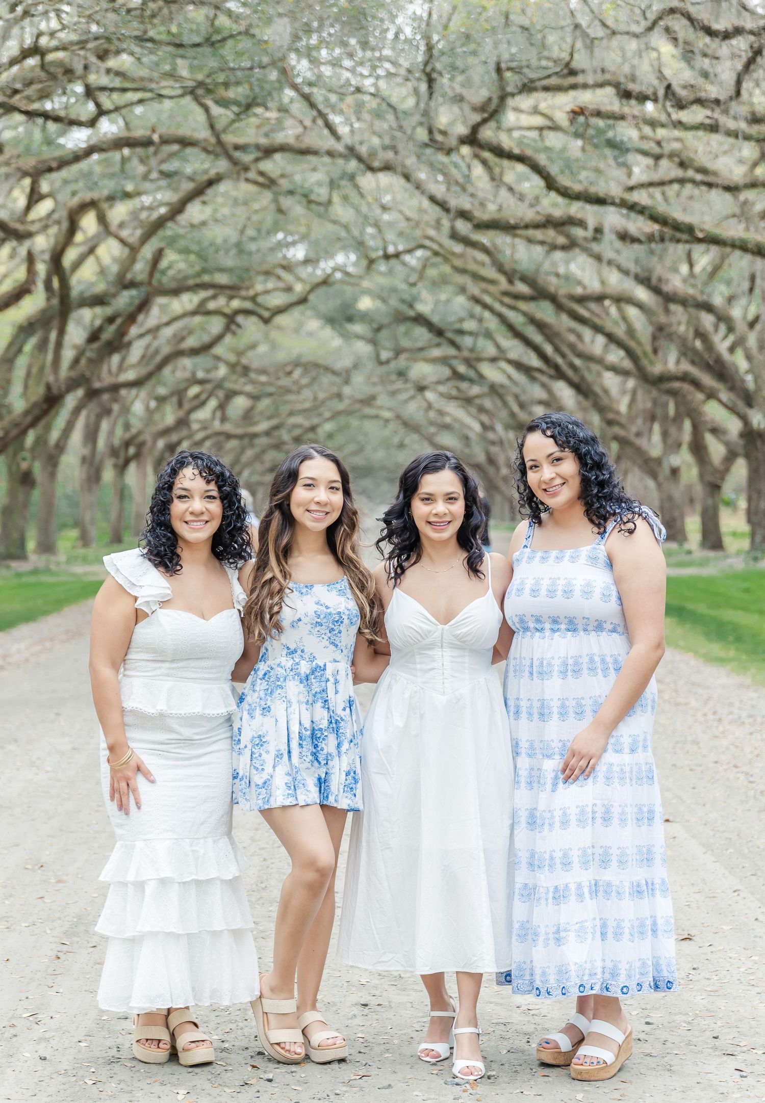 Four women in summery dresses posing together on a path beneath oak trees.