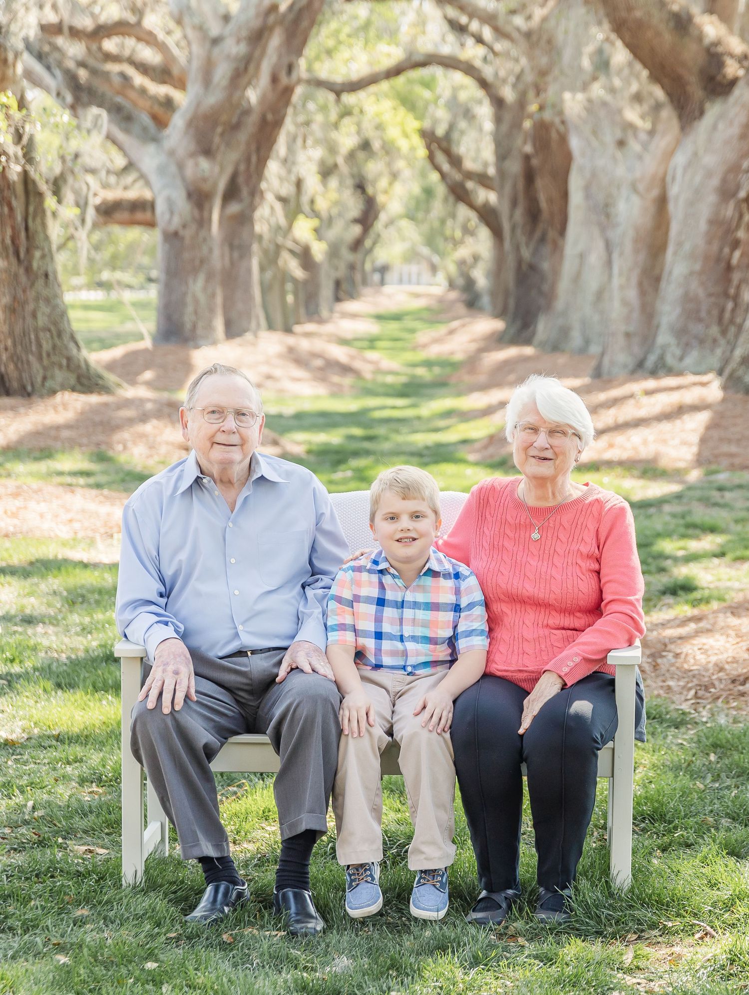 Three generations pose together on white bench beneath Spanish moss covered oak trees.