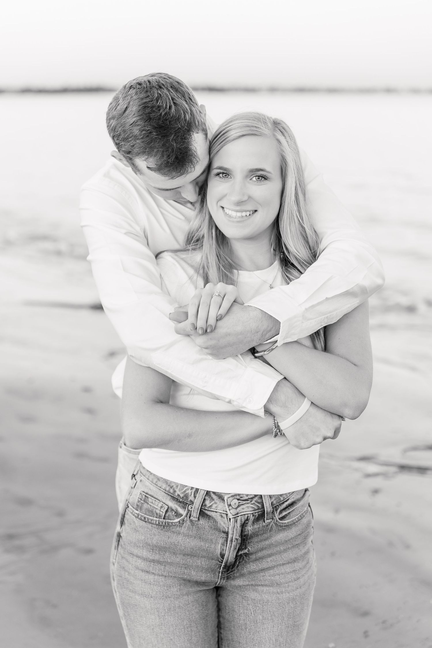 A couple embraces on a beach in casual white outfits during a romantic photoshoot.