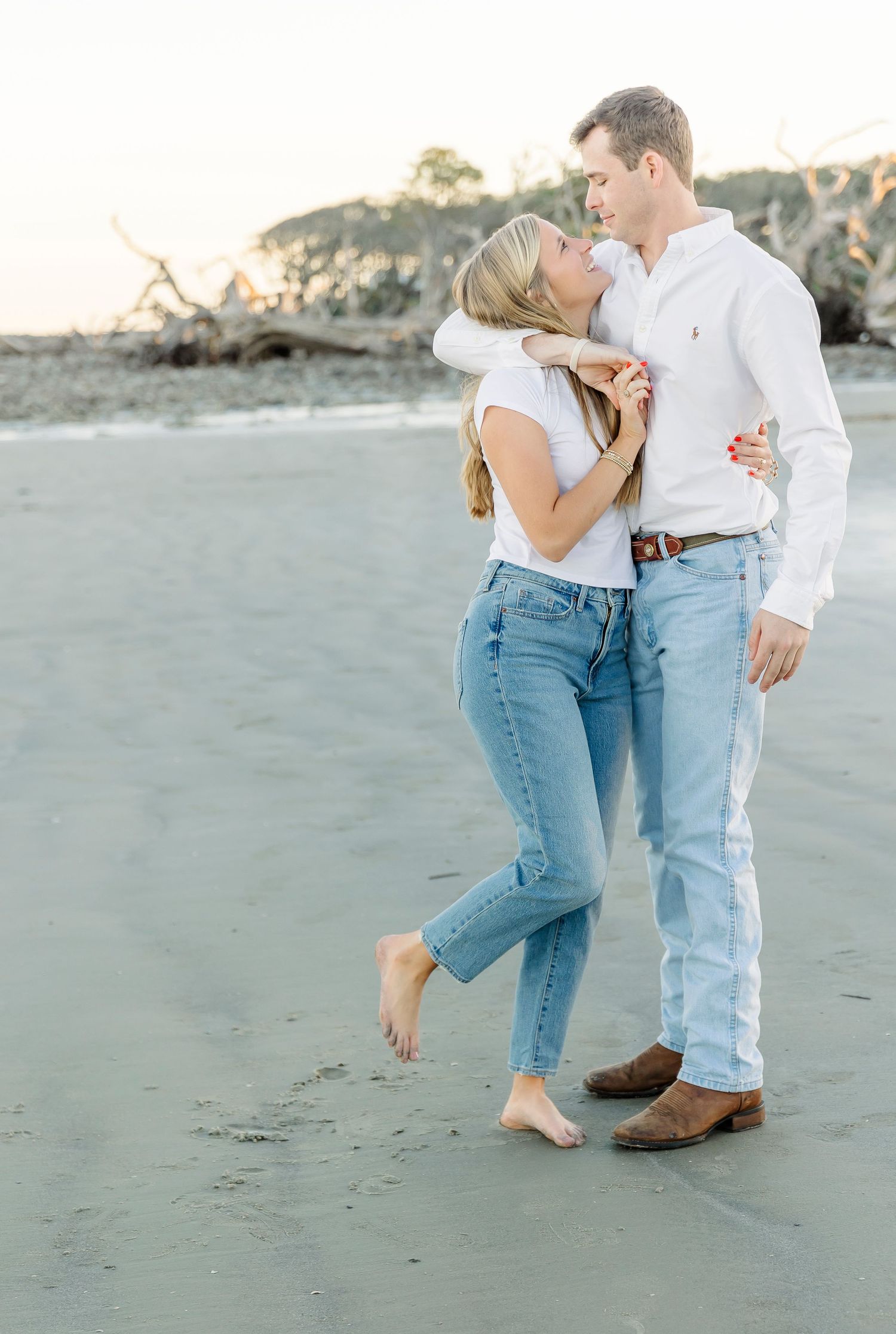Couple embracing on a beach at sunset wearing casual white and denim outfits.