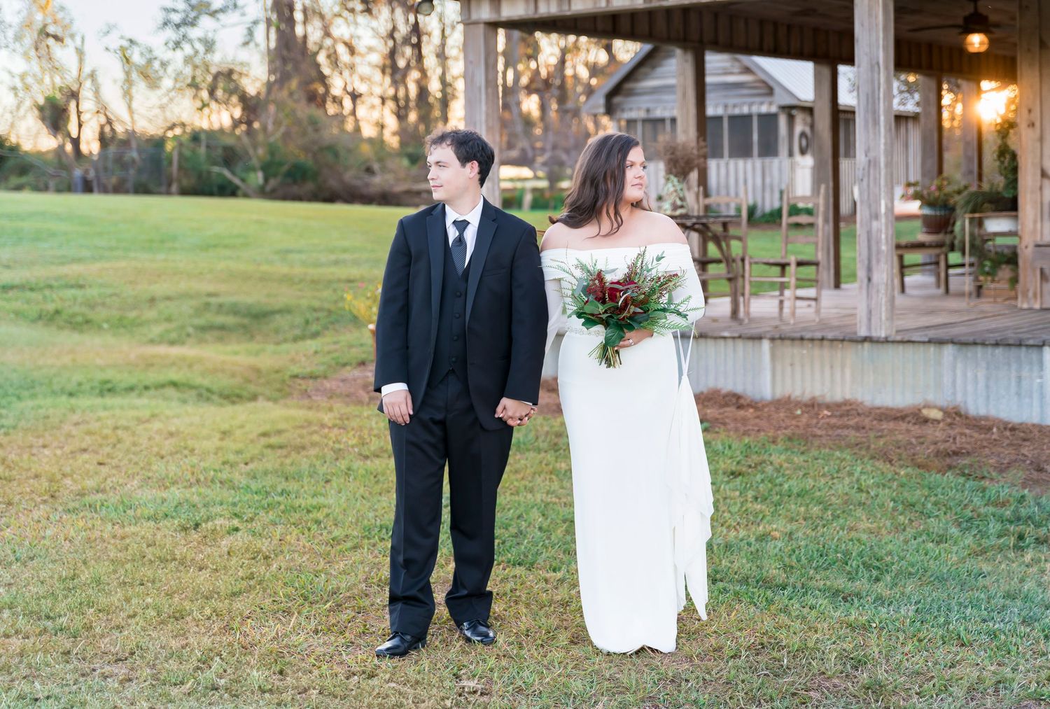 Wedding couple stands in front of a rustic pavilion during golden hour.