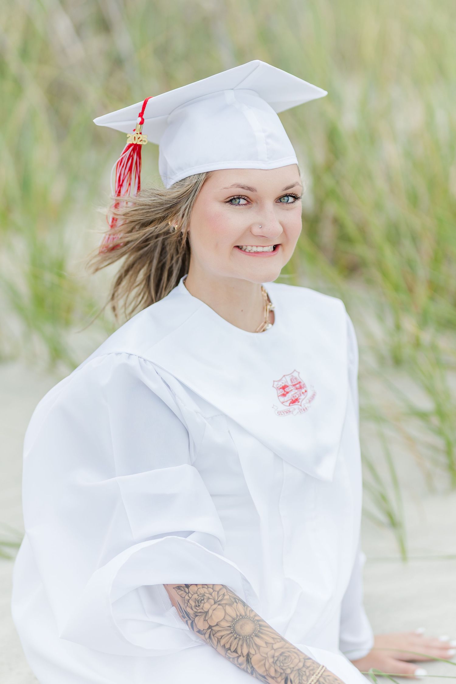 A graduate in white cap and gown poses outdoors among beach grass with visible tattoo sleeve.