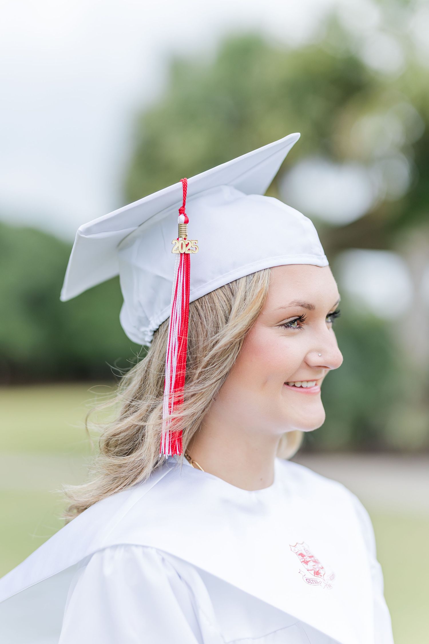 Graduate wearing a white cap and gown with red tassel smiles in an outdoor setting.