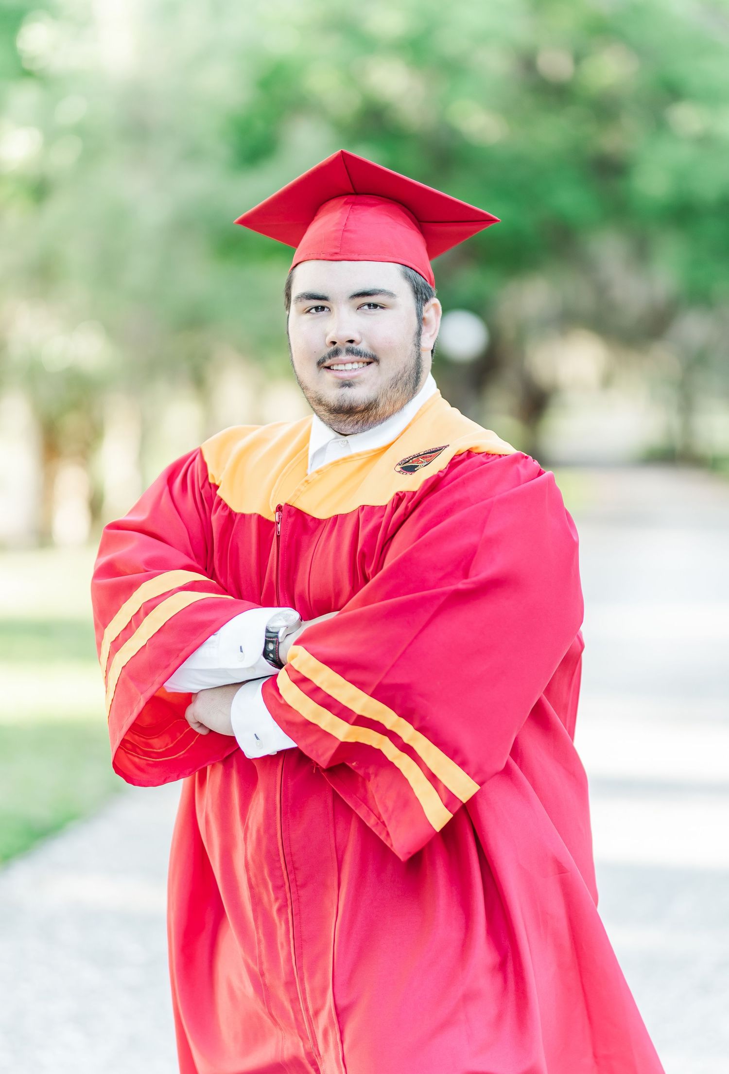 A graduate in red and gold graduation regalia poses outdoors with a blurred green background.
