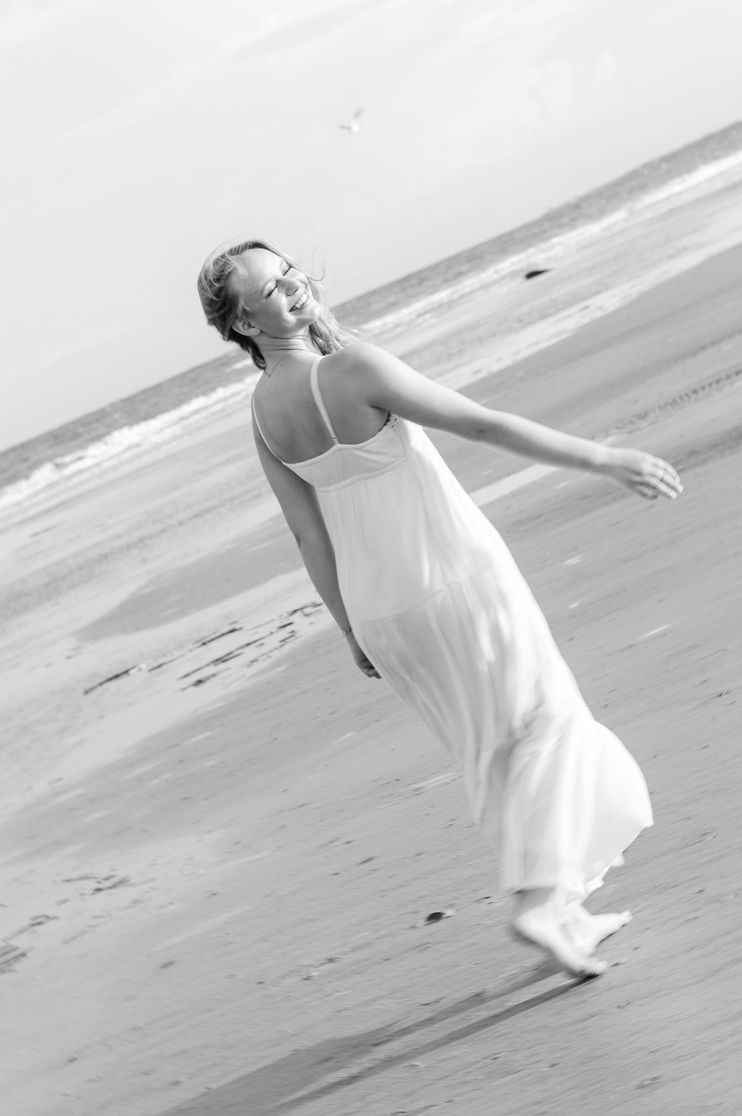 A graceful figure in a flowing white dress twirls joyfully on the beach in black and white.