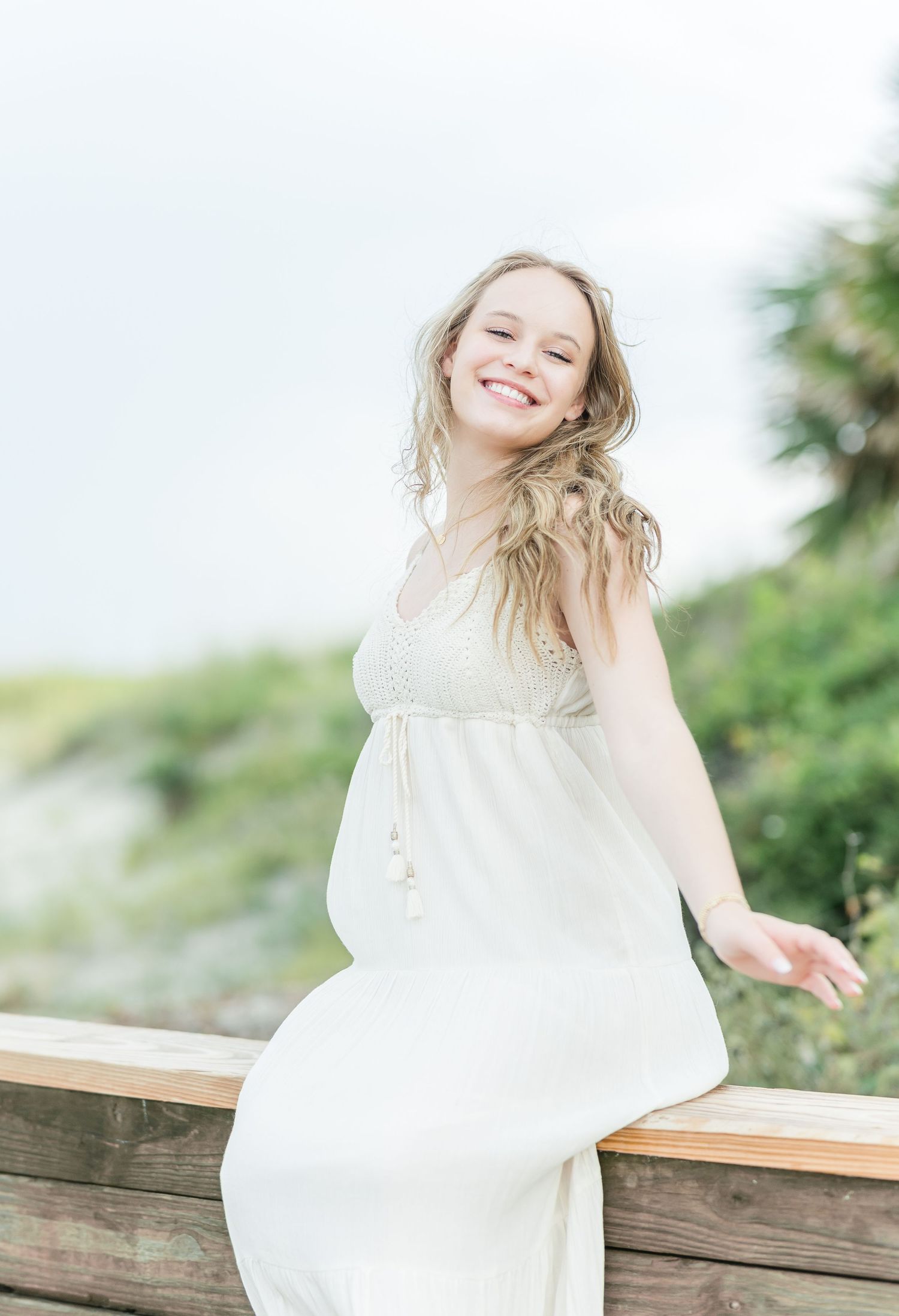 A woman in a flowing white dress poses on a wooden railing with greenery in the background.