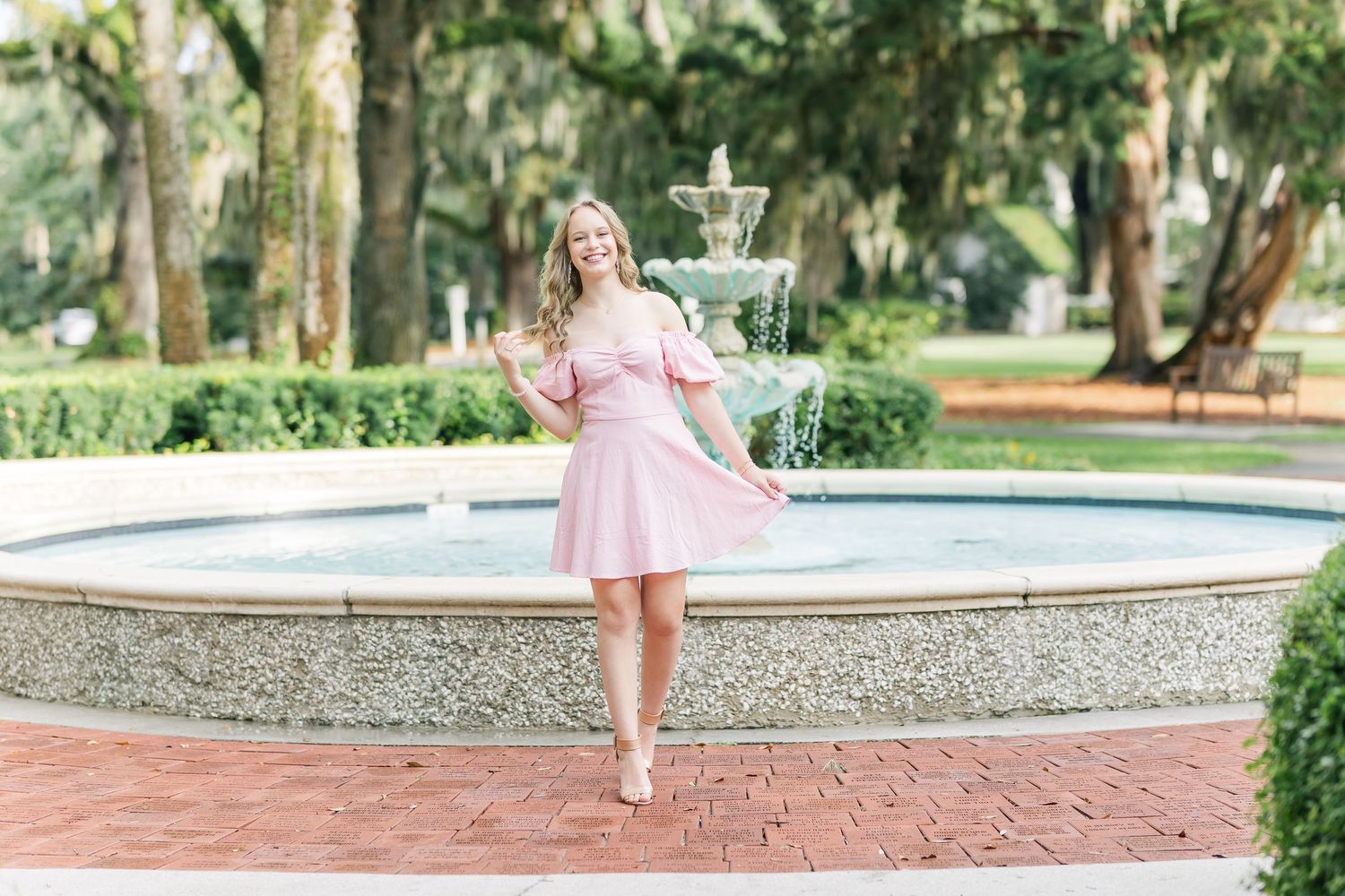 Someone in a pink dress twirls gracefully by a fountain in a lush garden setting with brick pathways.