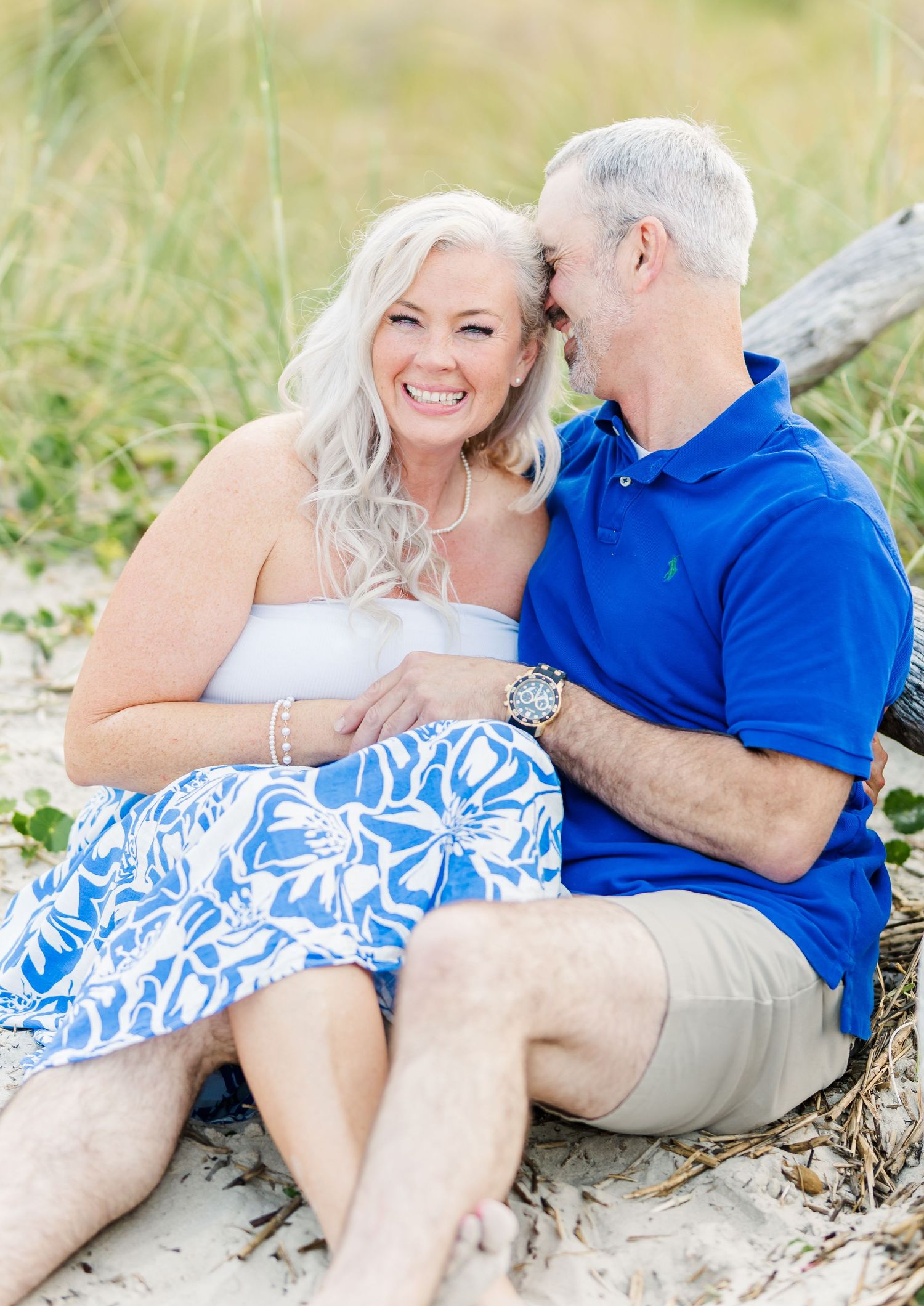 A loving couple in coordinating blue and white outfits share a joyful moment while sitting on a sandy beach.